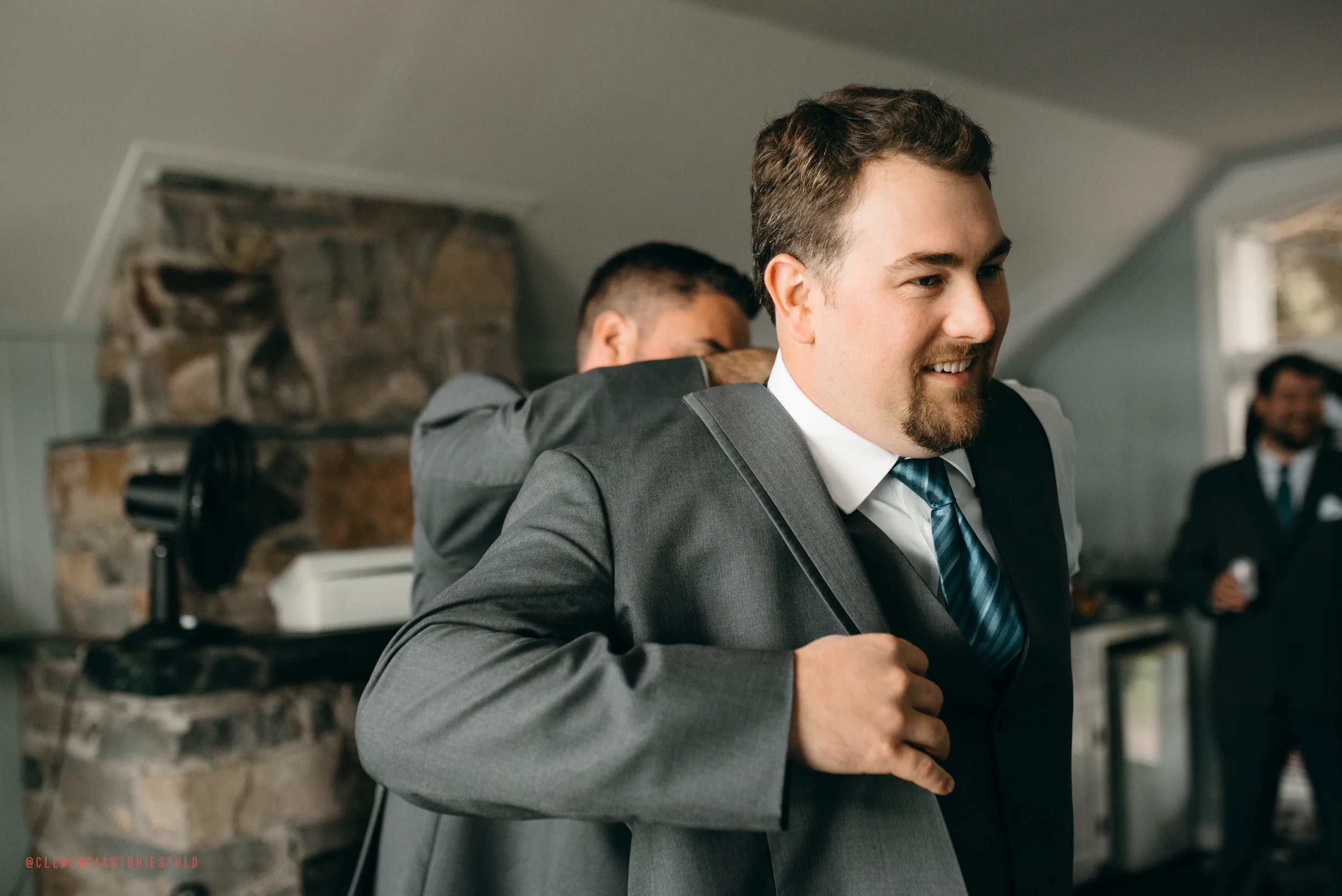 A man in a gray suit puts on a gray jacket while smiling, with two other men in suits in the background inside a room with a stone fireplace.