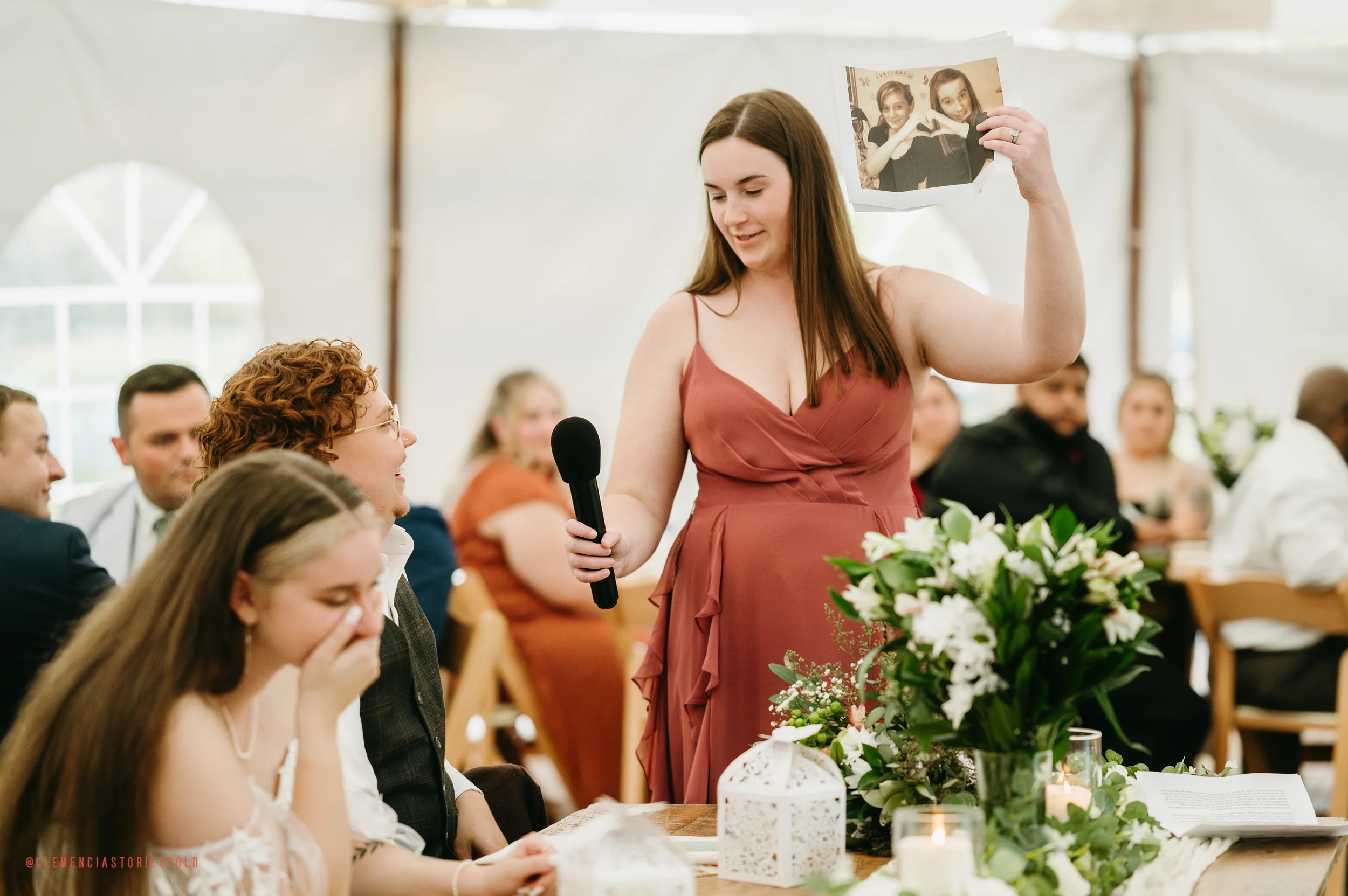A woman in a rust-colored dress holding a photo and a microphone at a wedding reception, with guests seated at a table and a large floral arrangement in front of her.