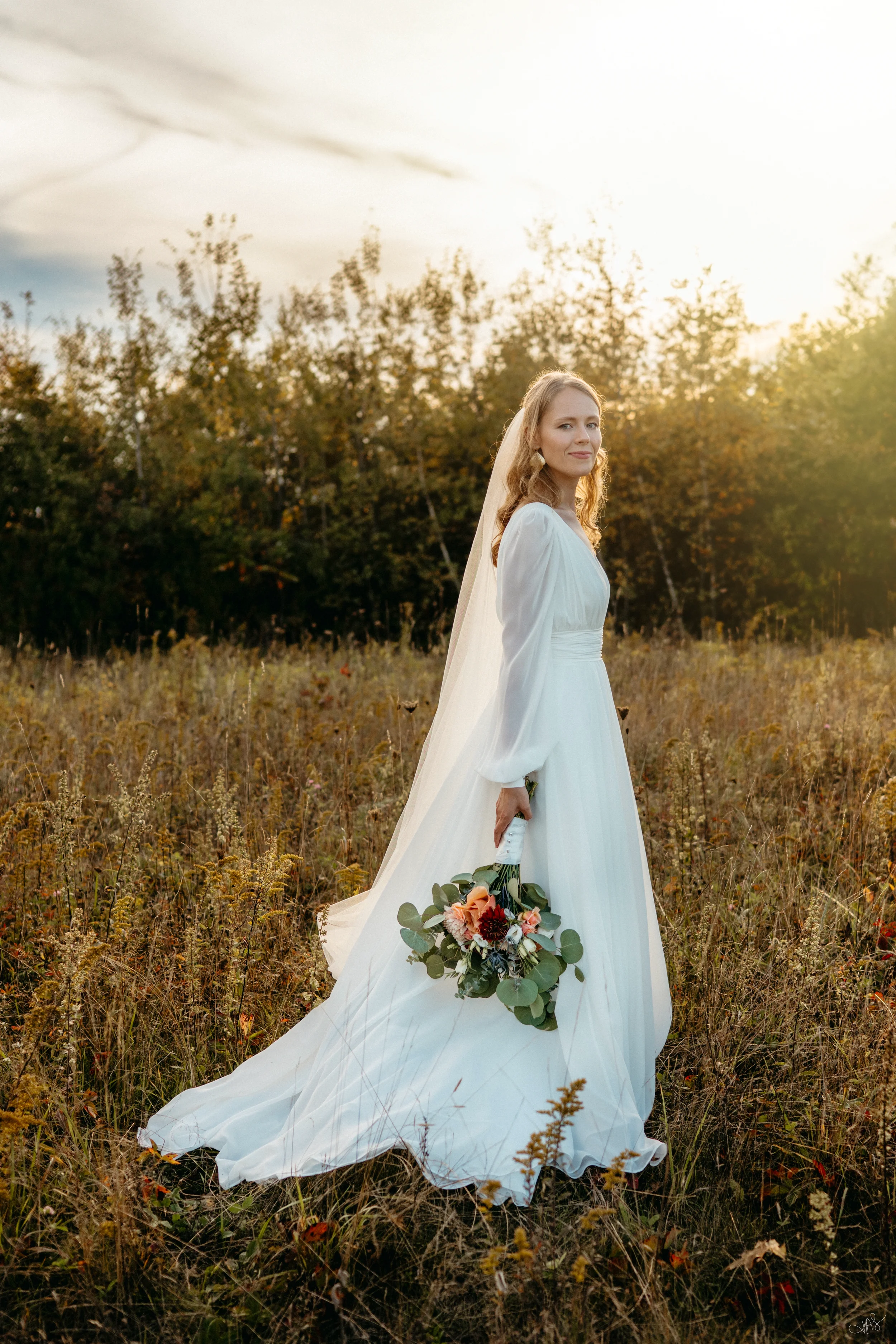 A bride in a white wedding dress and veil stands in a field of tall grass and wildflowers, holding a bouquet of flowers, with a backdrop of trees and a bright, backlit sky.