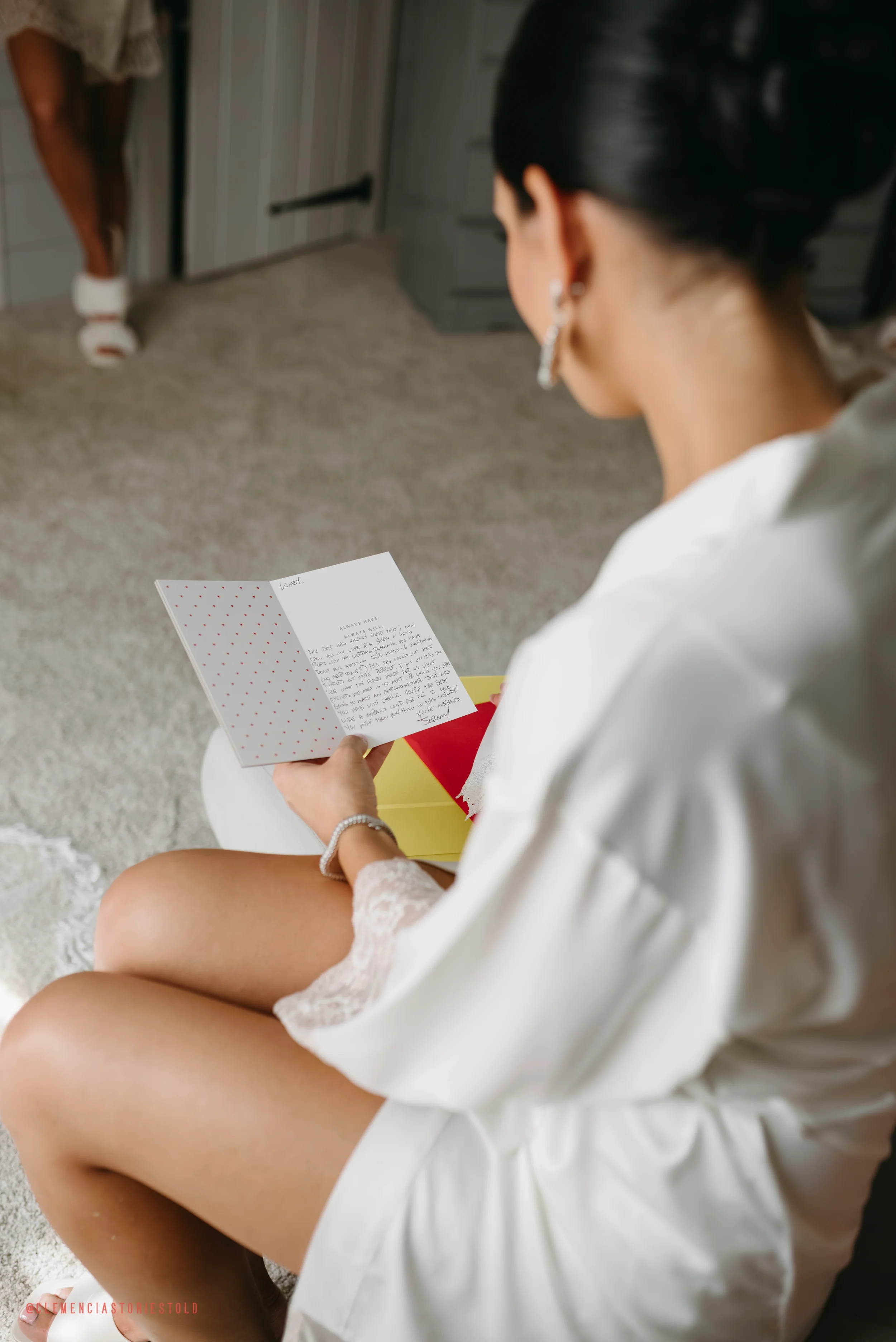 A woman in a white robe reading a card, seated with legs crossed, with a person in the background standing in a doorway.