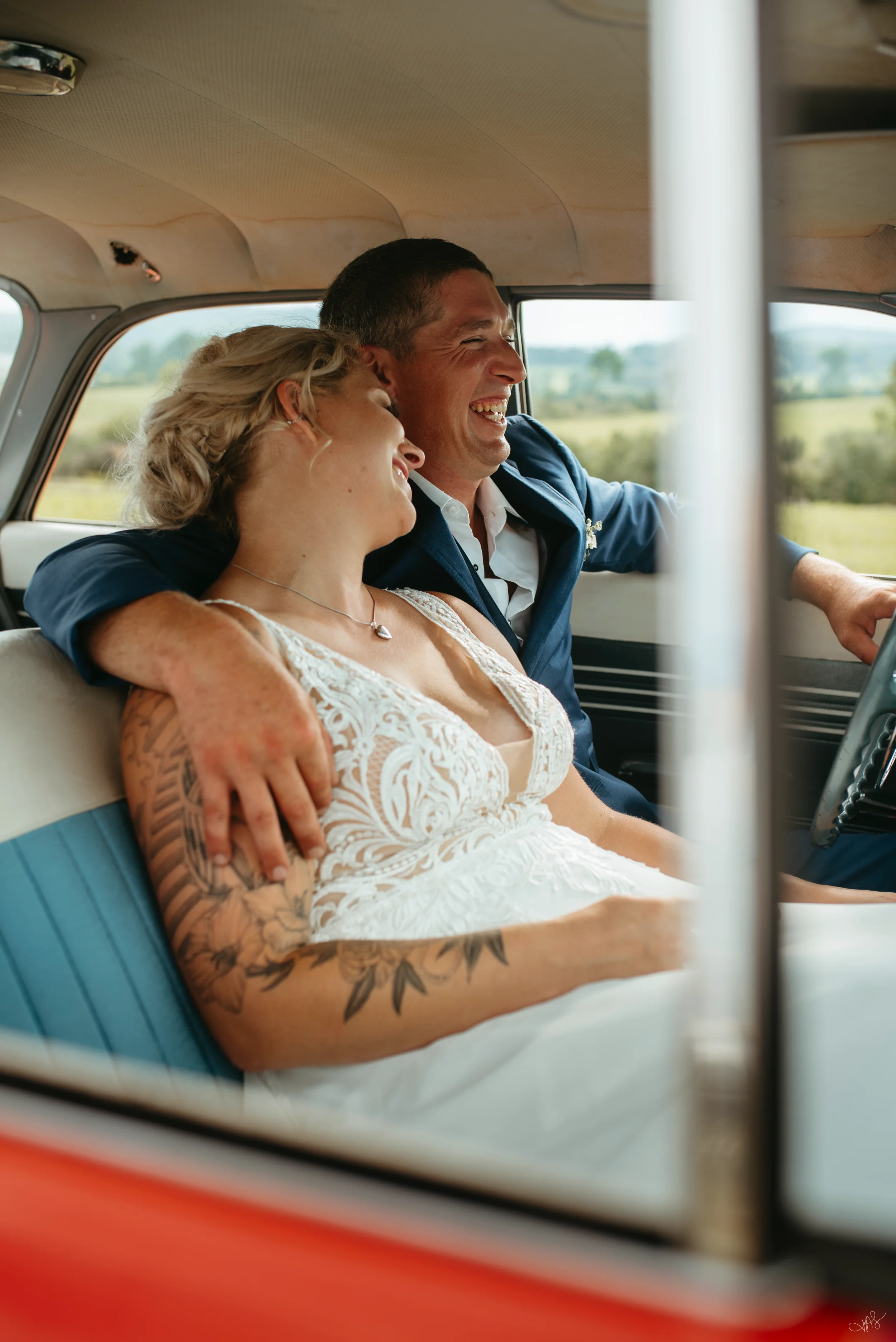 A couple sitting inside a vintage van, smiling and enjoying each other's company. The woman has blonde hair and visible tattoos on her arm, while the man has short dark hair and is wearing a blue jacket over a white shirt. They are sitting close, with the woman leaning her head on the man's shoulder as they share a joyful moment, with a scenic landscape visible through the windows.