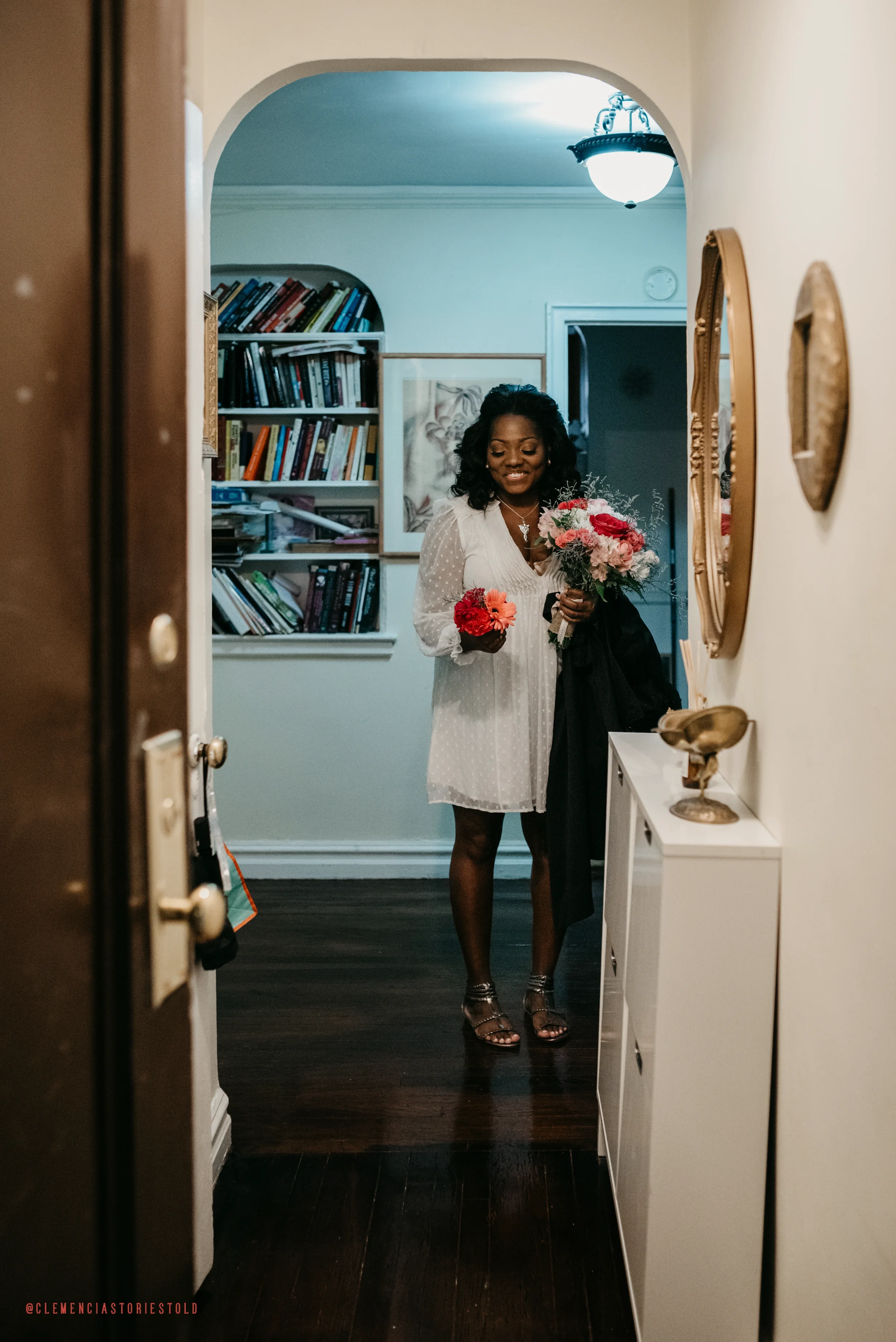 A woman standing in a hallway holding a bouquet of pink and red flowers, smiling.