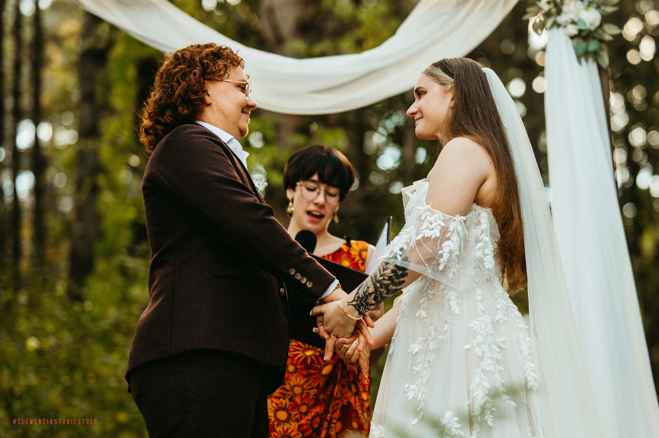 A same-sex couple getting married outdoors, holding hands and smiling at each other during the wedding ceremony beneath a white draped arch in a wooded area, with an officiant reading from a book in the background.