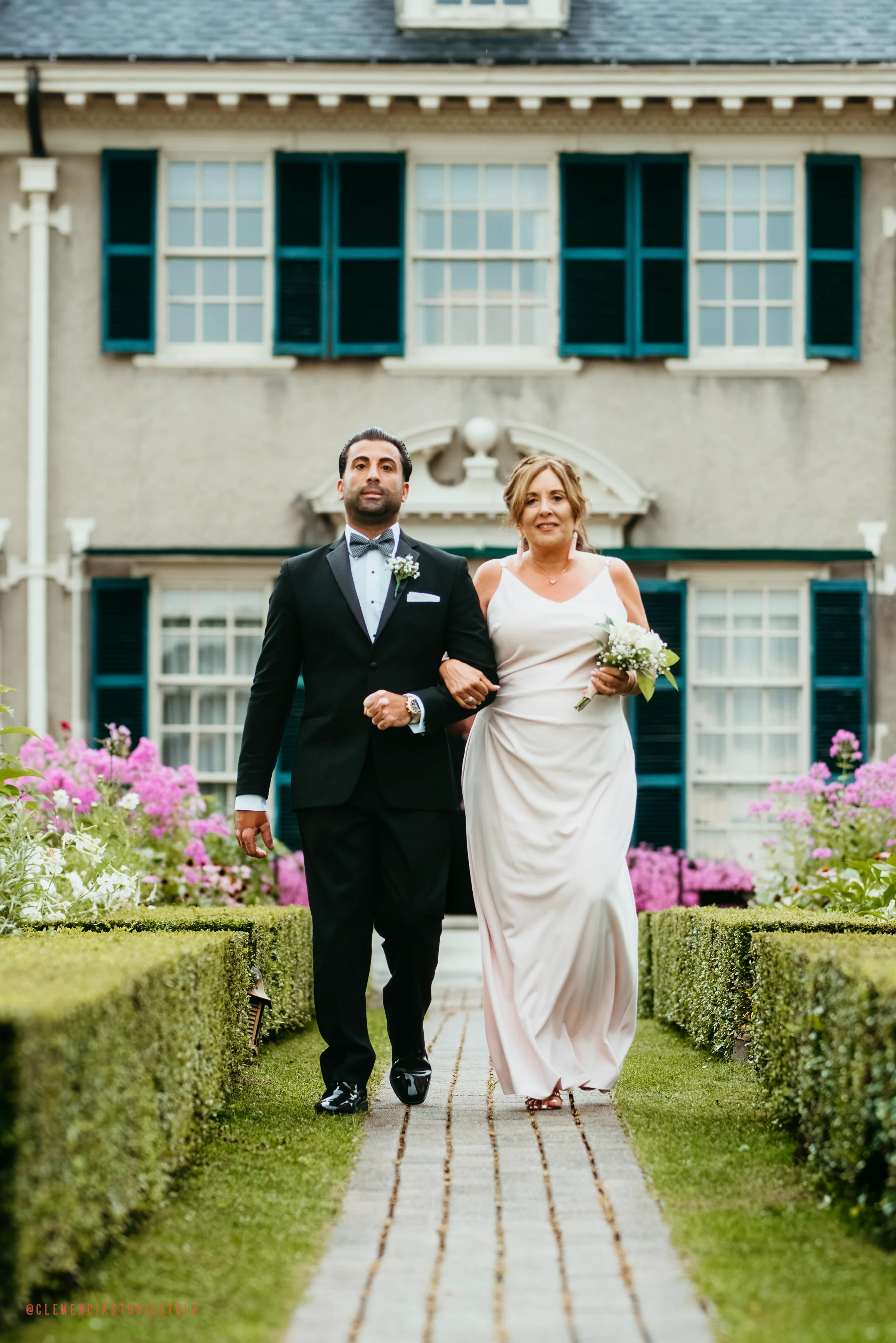 A bride and groom walking arm in arm down a garden path in front of a large house with blue shutters and pink flowers.