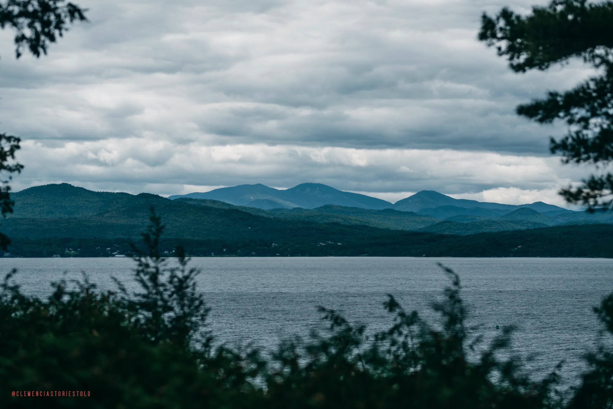 Scenic view of a lake surrounded by tree silhouettes in the foreground and a mountain range with cloudy sky in the background.