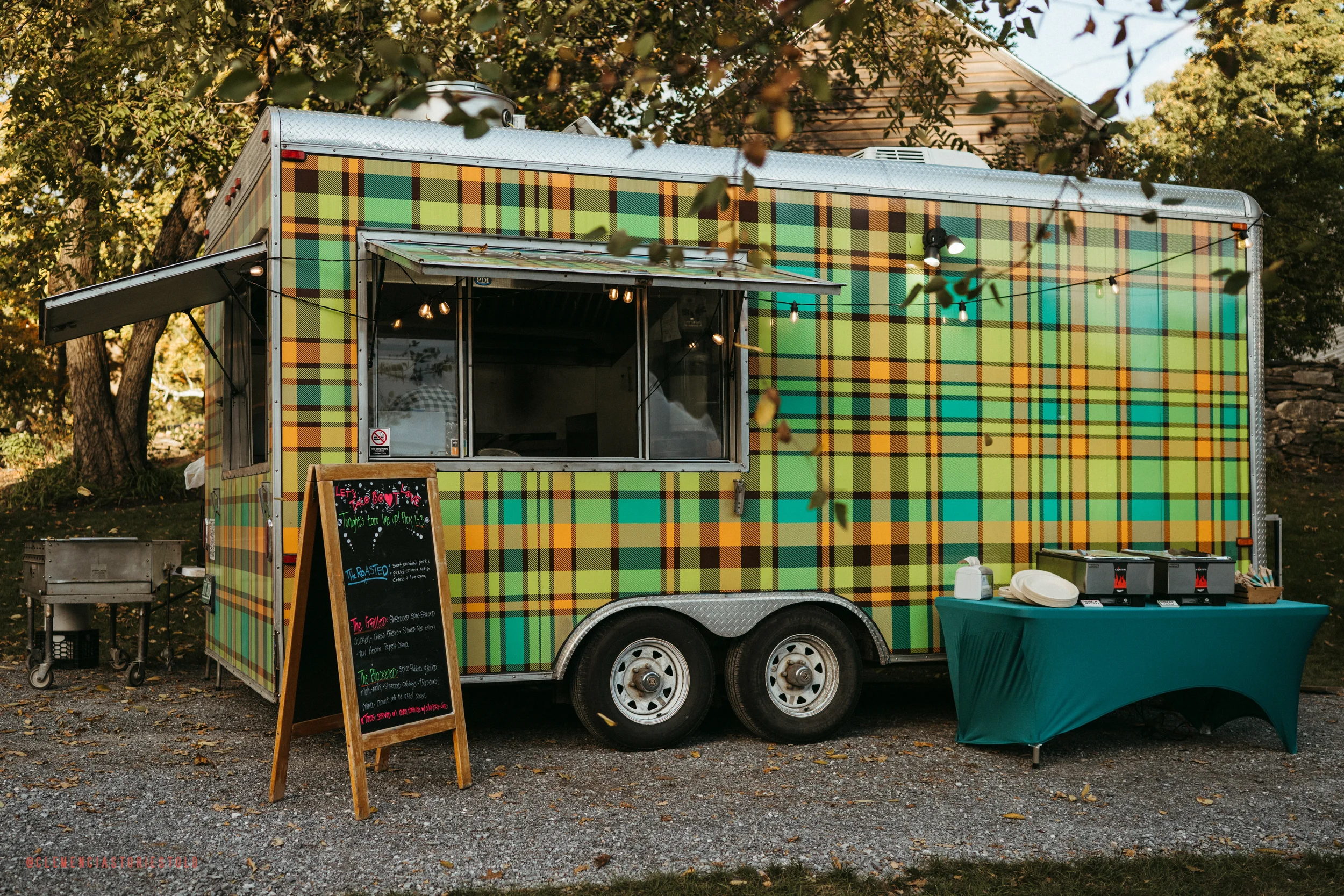 A colorful food truck with a green, yellow, and orange plaid pattern, set outdoors on a gravel surface with trees and a house in the background. It has a window open for service, a chalkboard menu sign in front, and a table with containers of condime