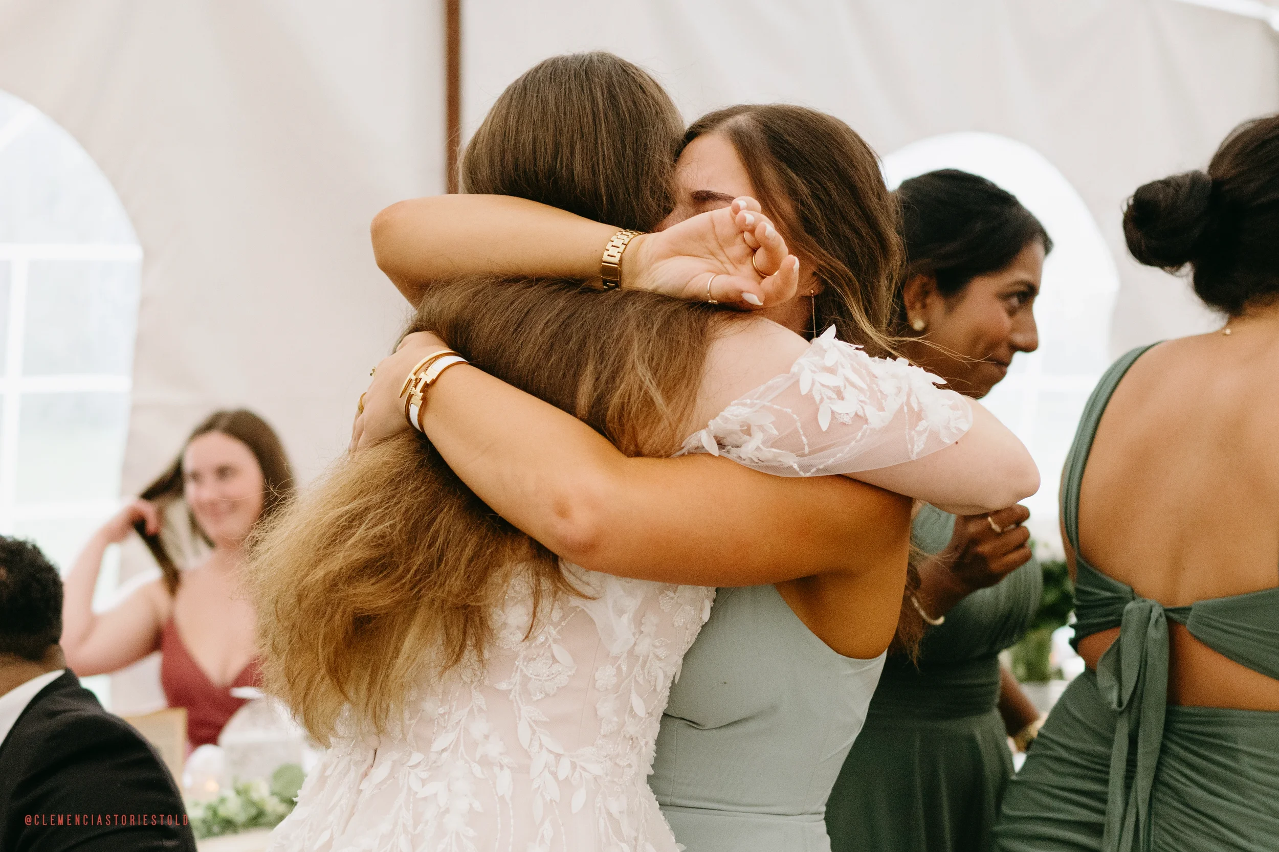 Two women hugging, one in a white dress and the other in a light green dress, at a social gathering or celebration, with people in the background.