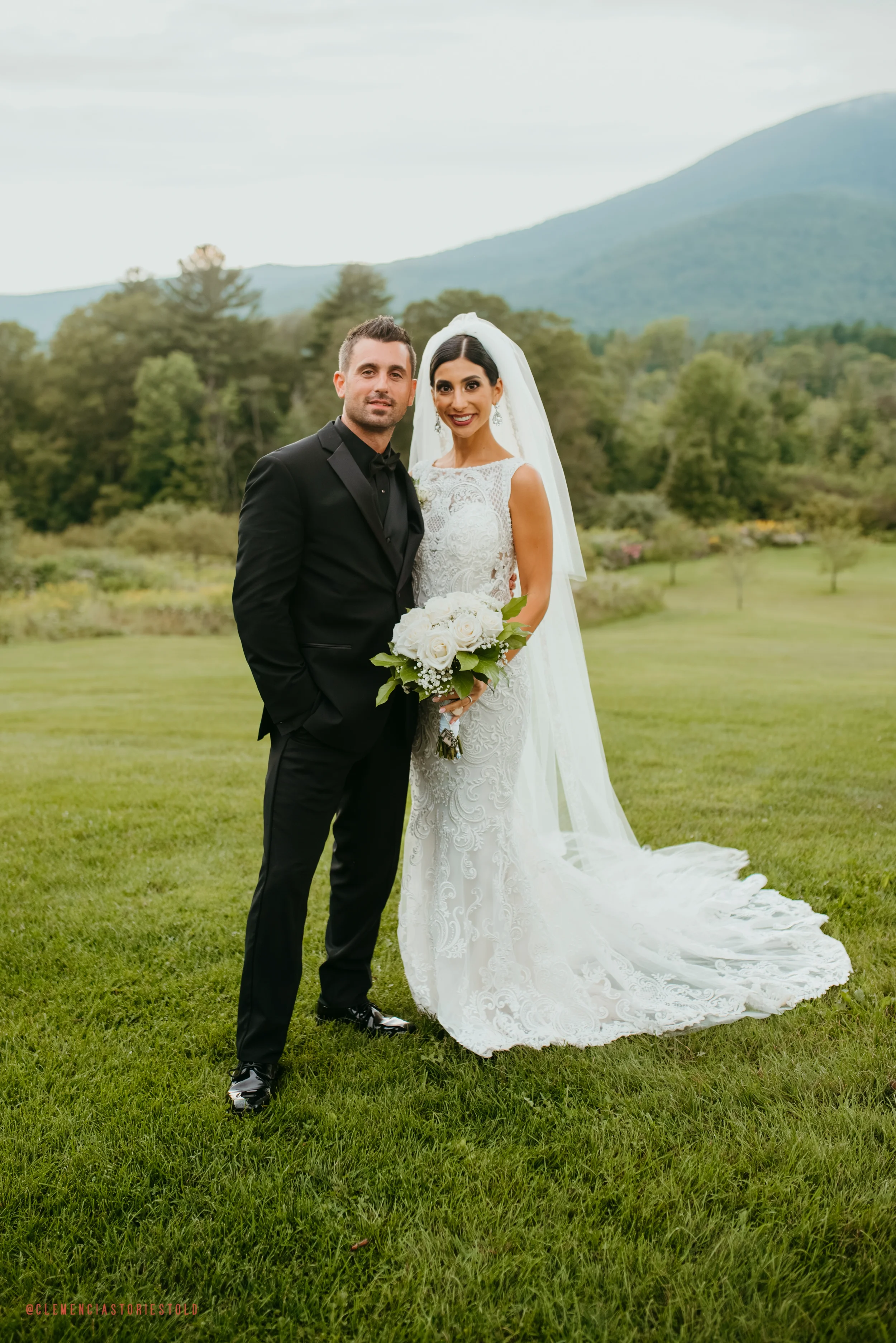 A bride and groom standing together on a grassy field with trees and mountains in the background. The bride is wearing a white wedding dress and veil, holding a bouquet of white roses. The groom is dressed in a black tuxedo.