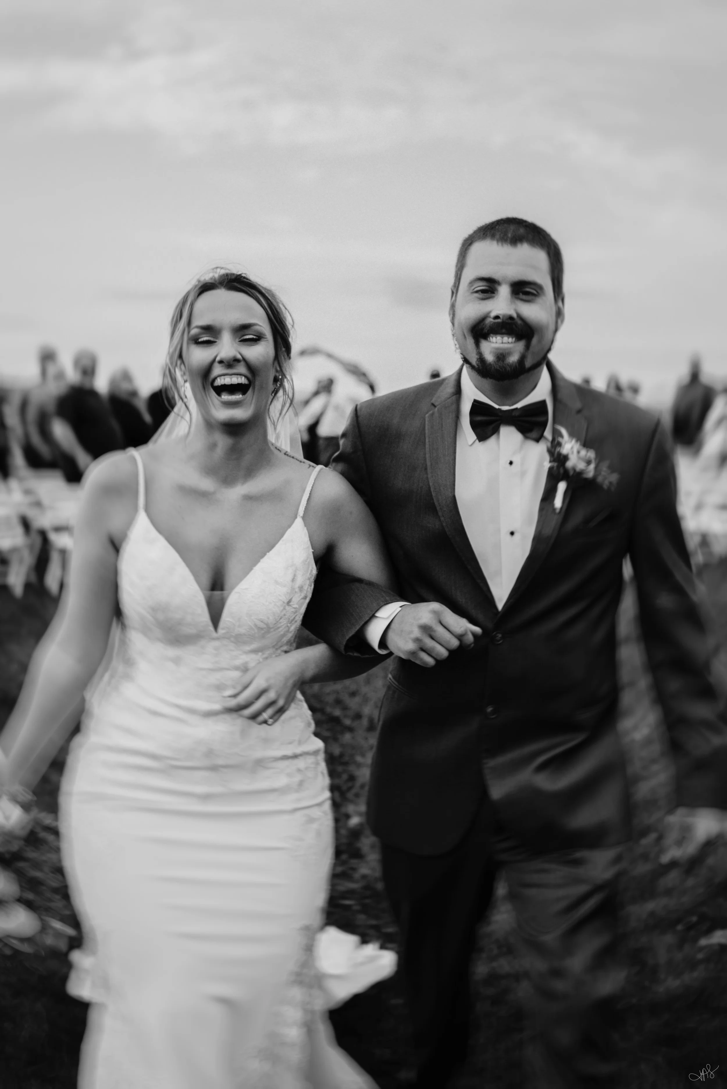 Black and white photo of a smiling couple at their wedding, walking outdoors with arm linked, surrounded by wedding guests and a cloudy sky.