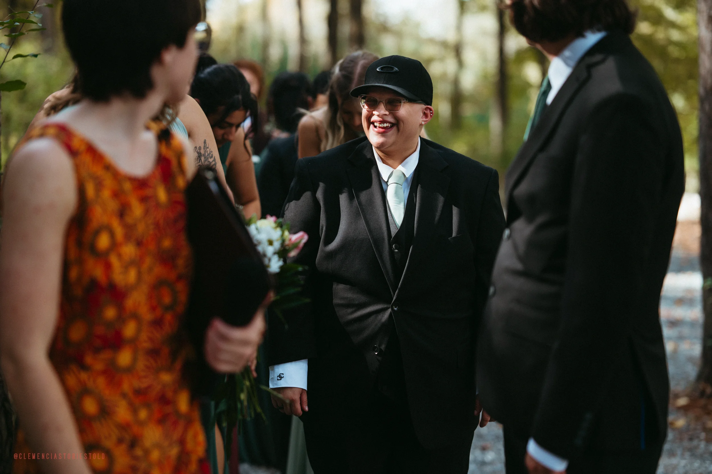 People in formal attire gathered outdoors, smiling, with a man in the center wearing a black suit, white shirt, and glasses, engaging with others during a wedding or celebration.