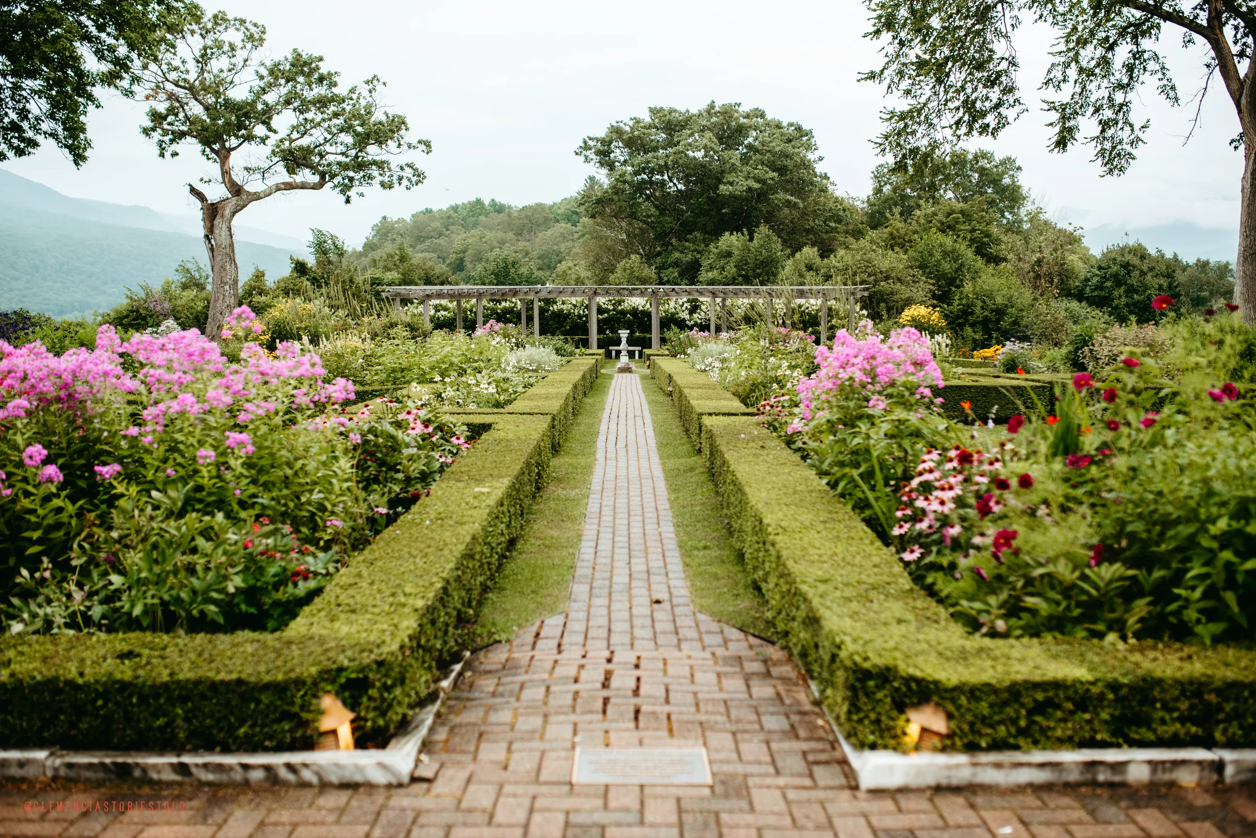 A paved garden pathway flanked by well-manicured green hedges and blooming flowers leads toward a fountain and a wooden pergola in the distance, with trees and mountains in the background.