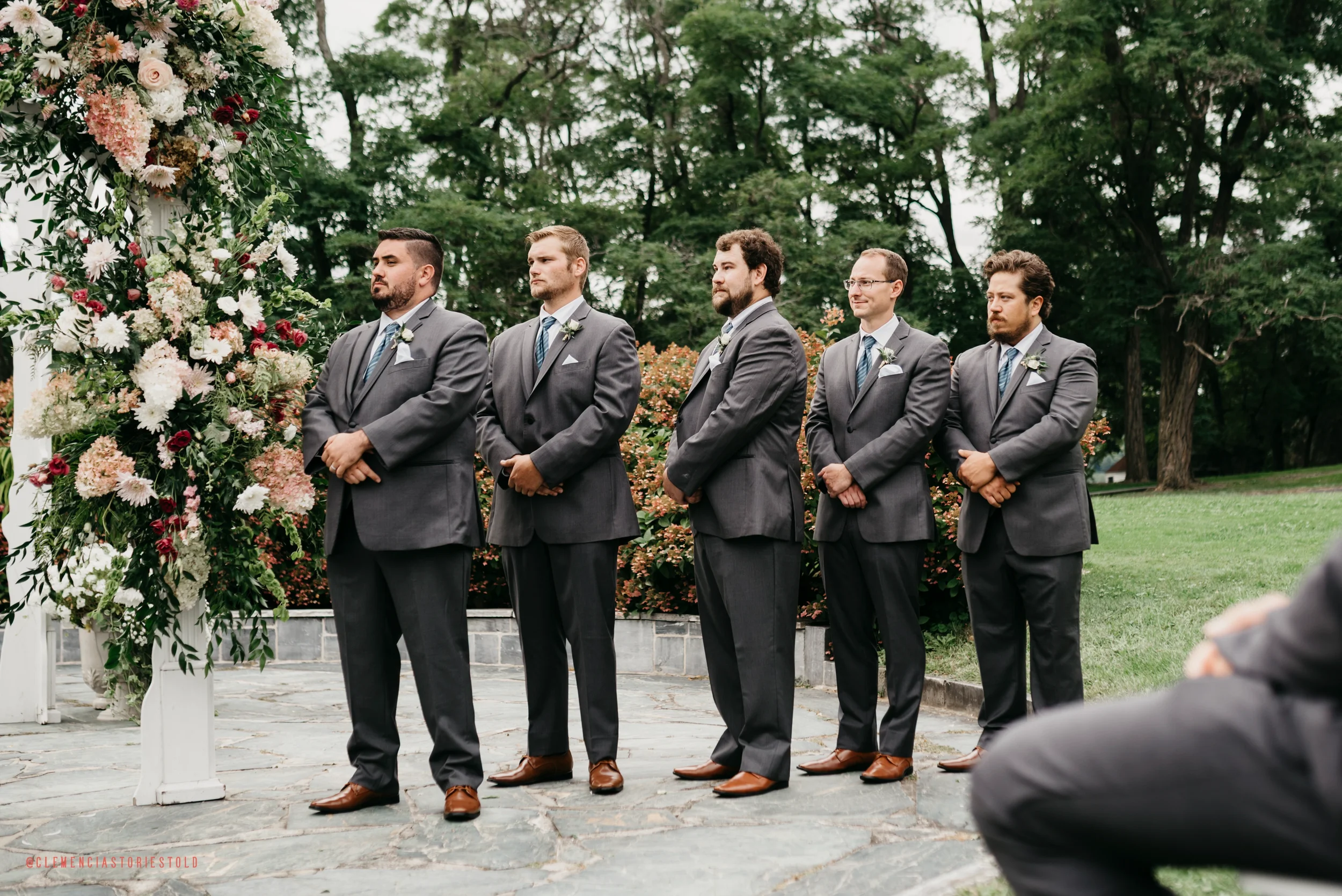 Five men dressed in gray suits standing in line outside during a wedding ceremony, facing forward with hands clasped in front, near a floral arch decorated with pink, white, and red flowers, with trees and bushes in the background.