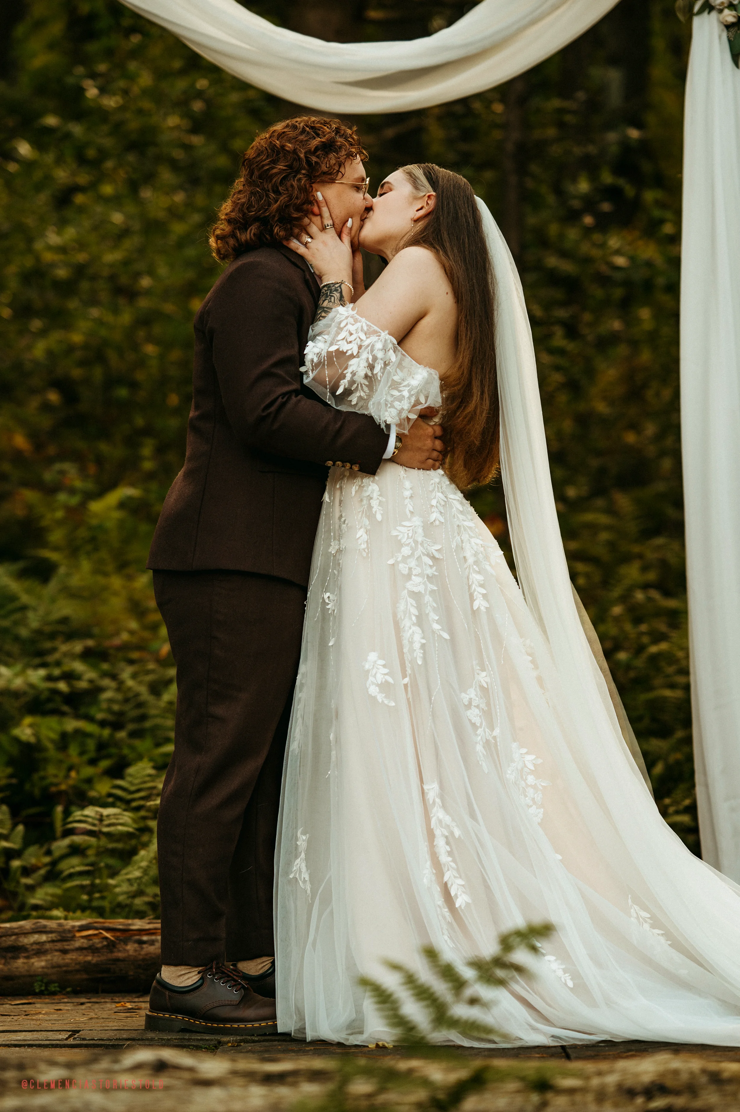 A bride and bride kiss during their wedding ceremony outdoors, with a natural background and white fabric decorations.