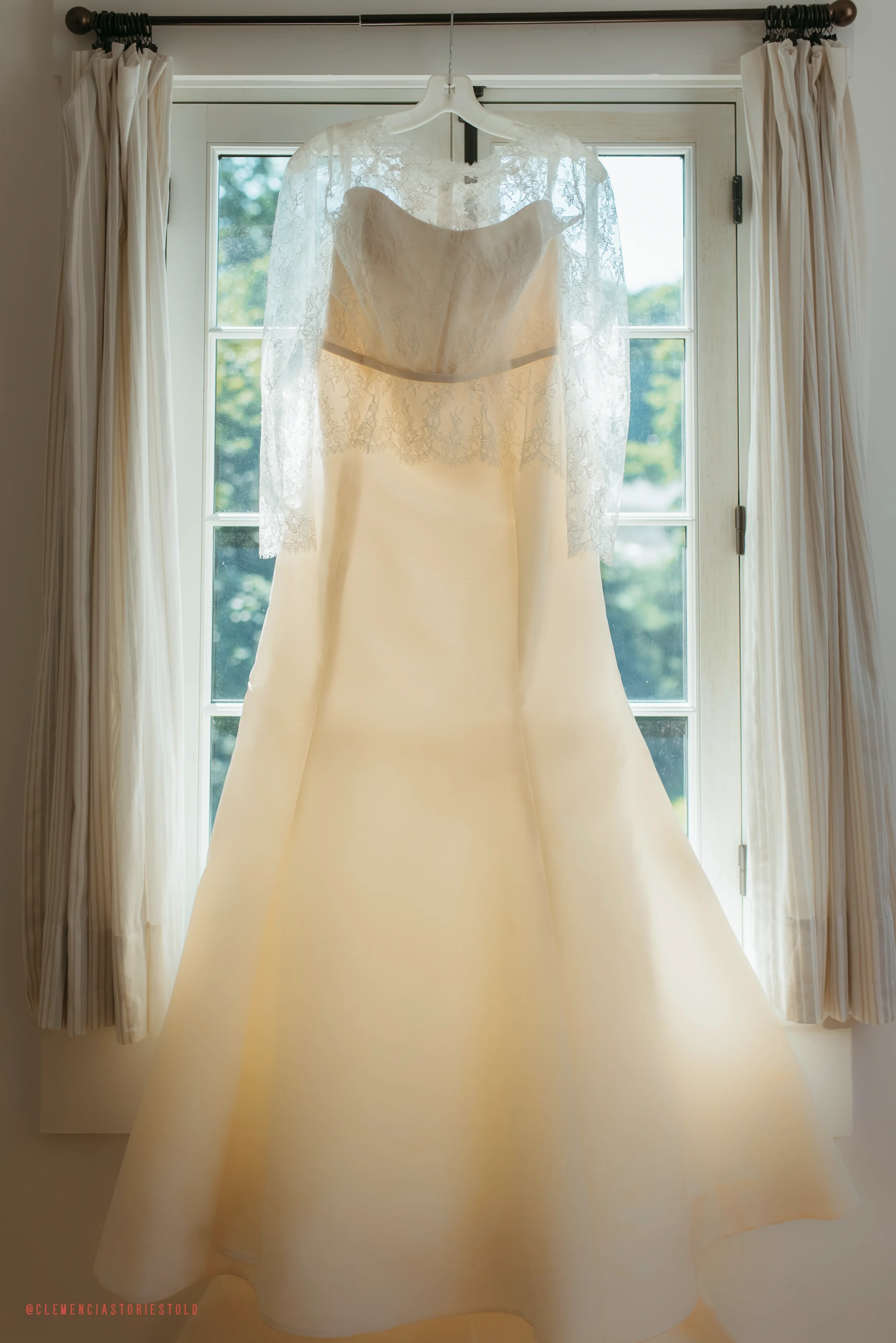 A wedding dress hanging on a white hanger in front of a glass door, with lace overlay on the top and sheer lace sleeves, illuminated by natural light.