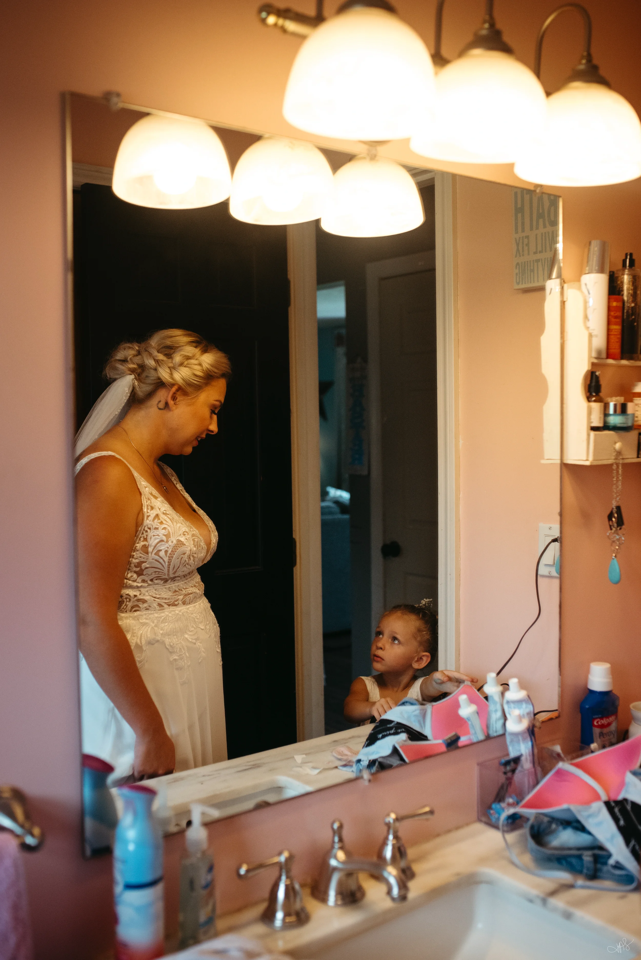 A woman in a wedding dress looking down at a young girl in a bathroom. The bathroom mirror reflects overhead lights and a sign that says 'BATH' and 'VITAMIN'. The countertop has toiletries and a pink bag, and the scene is warmly lit.