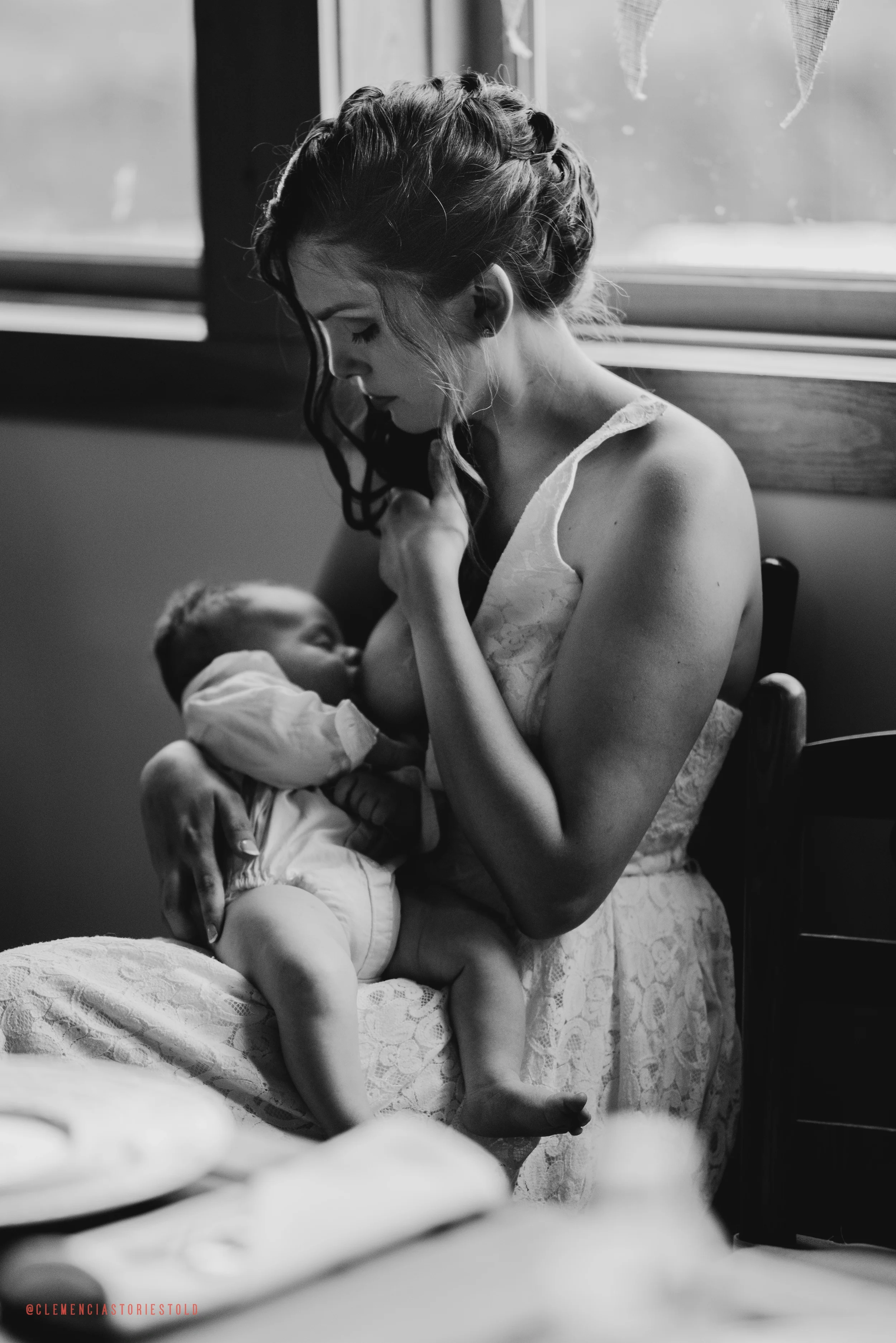 A woman with wavy hair holds a sleeping baby on her lap, sitting by a window in black and white.