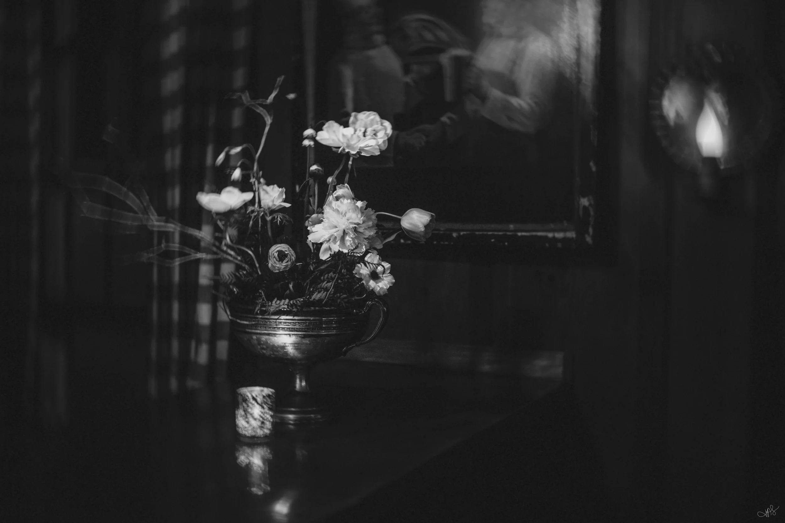 A black and white photograph of a floral arrangement in a decorative metal vase, placed on a wooden surface. In the background, there is a reflection in a mirror showing a person holding a camera.