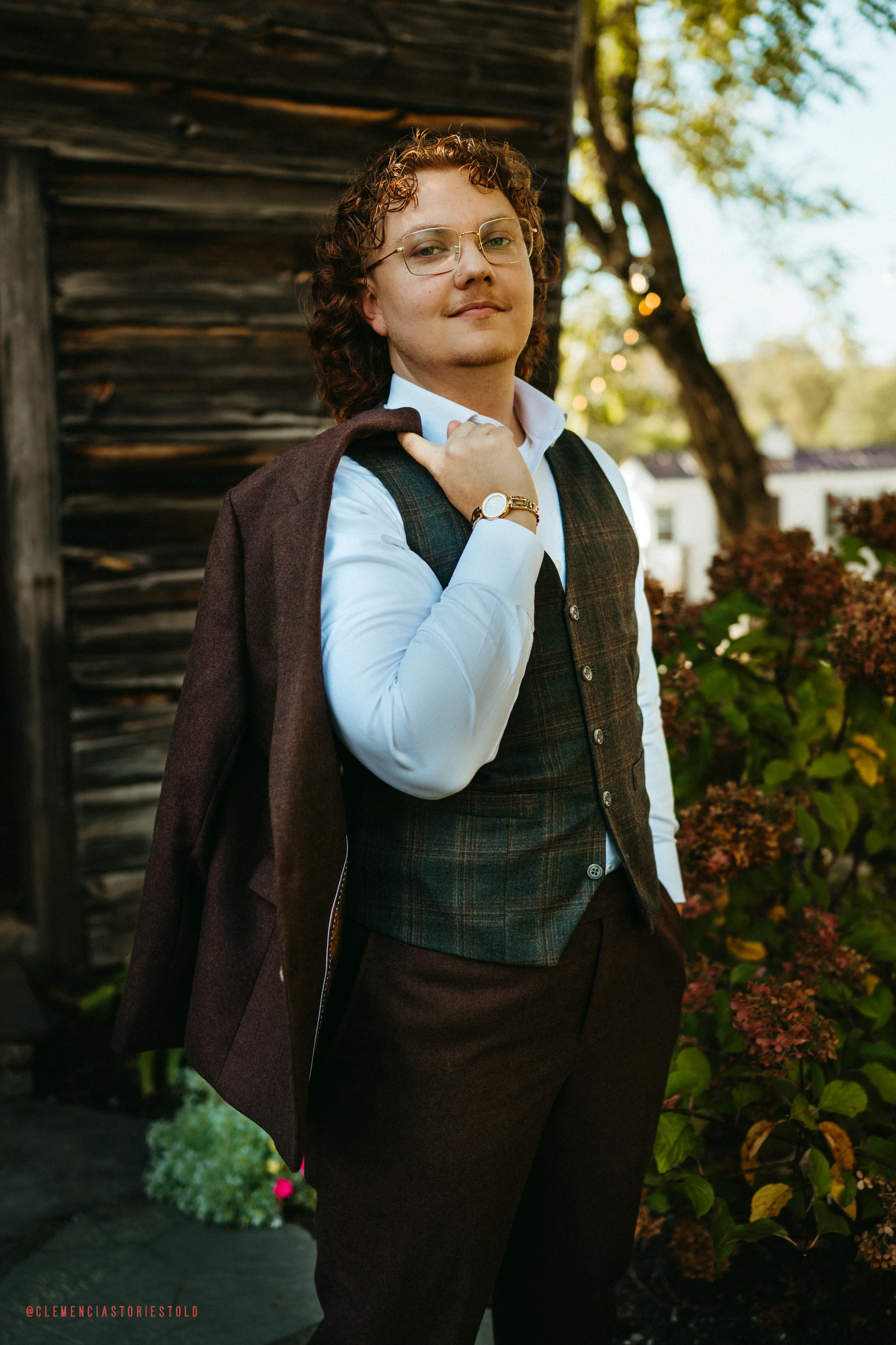 A person with curly hair wearing glasses, a white shirt, and a plaid vest, carrying a brown coat over their shoulder, standing outdoors near greenery and a wooden structure.