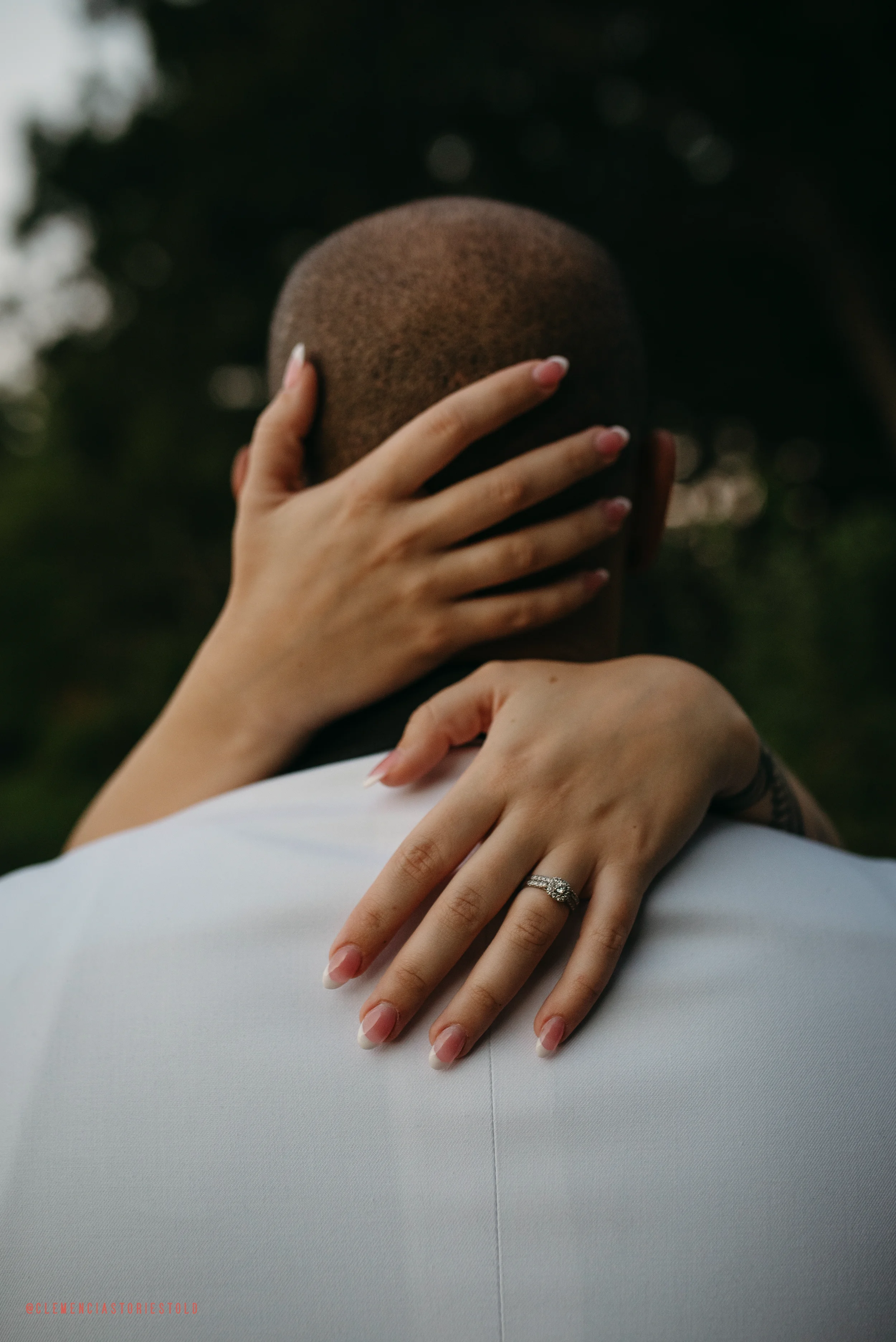 A person with a shaved head is being embraced from behind by a woman, with her arms wrapped around their head and shoulders. The woman is wearing a wedding ring on her left hand, which rests on the person's shoulder. The background is outdoors with blurred trees.