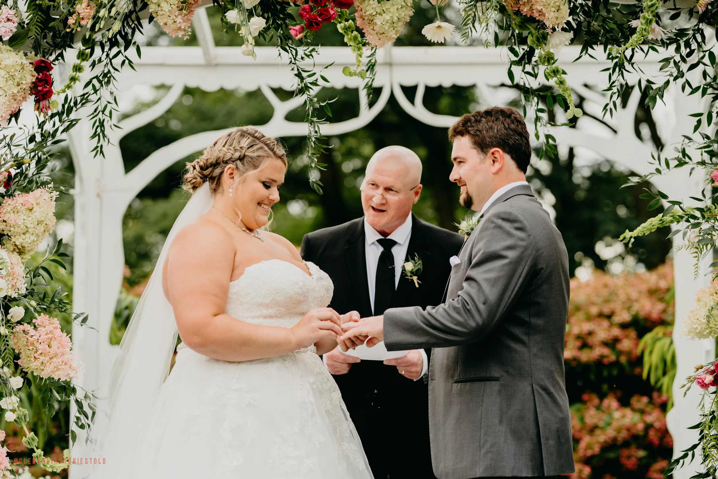 A bride and groom exchanging rings during their outdoor wedding ceremony with officiant present, under a floral arch.