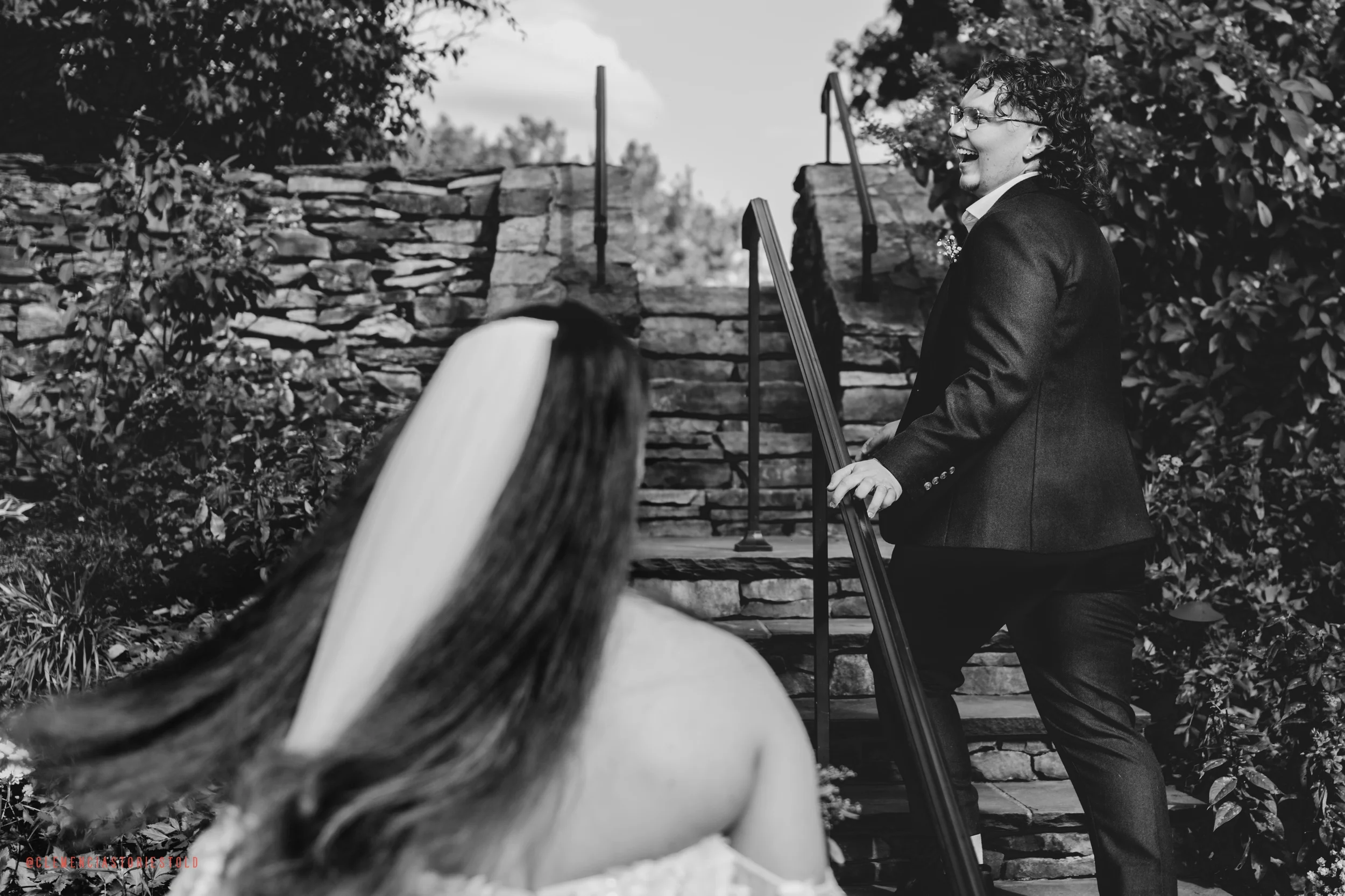 A black and white photo of a wedding couple outdoors, with the groom standing on stairs holding a guitar and laughing, while the bride sits in the foreground with her back to the camera.