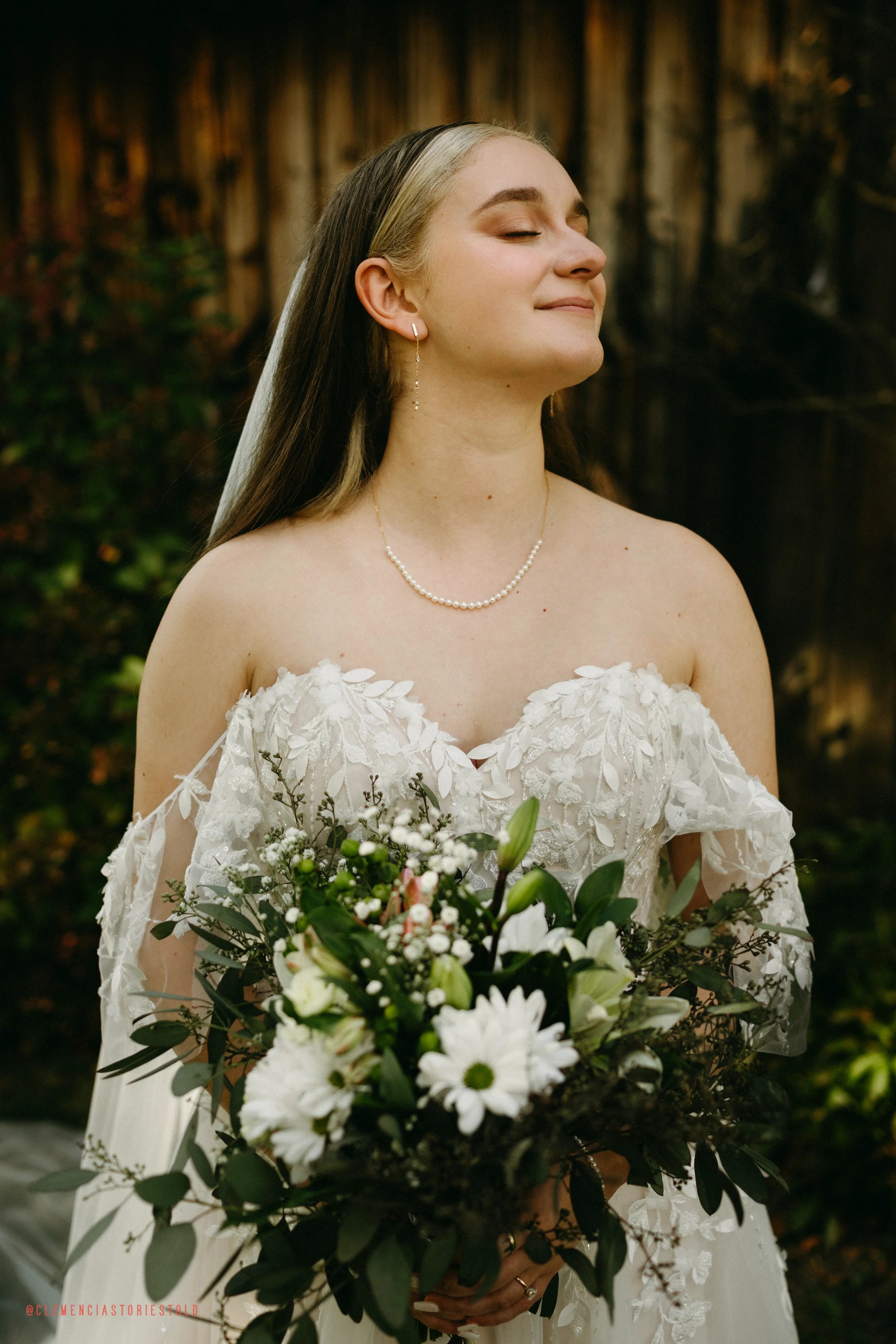 A bride wearing a white wedding dress with floral lace details, holding a bouquet of white daisies and lilies, standing outdoors with a wooden fence in the background, eyes closed and smiling softly.
