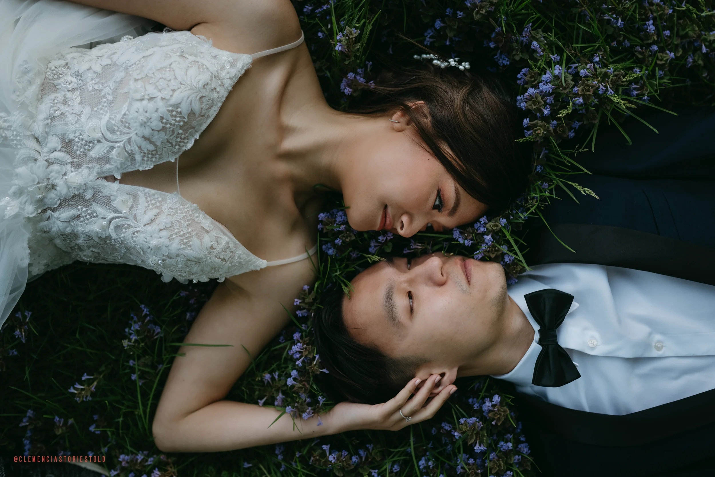 A woman and a man lying on the grass surrounded by small purple flowers. The woman is wearing a white lace dress, and the man is dressed in a white shirt with a black bow tie. They are facing each other with their heads close together, and the woman is gently holding the man's head.