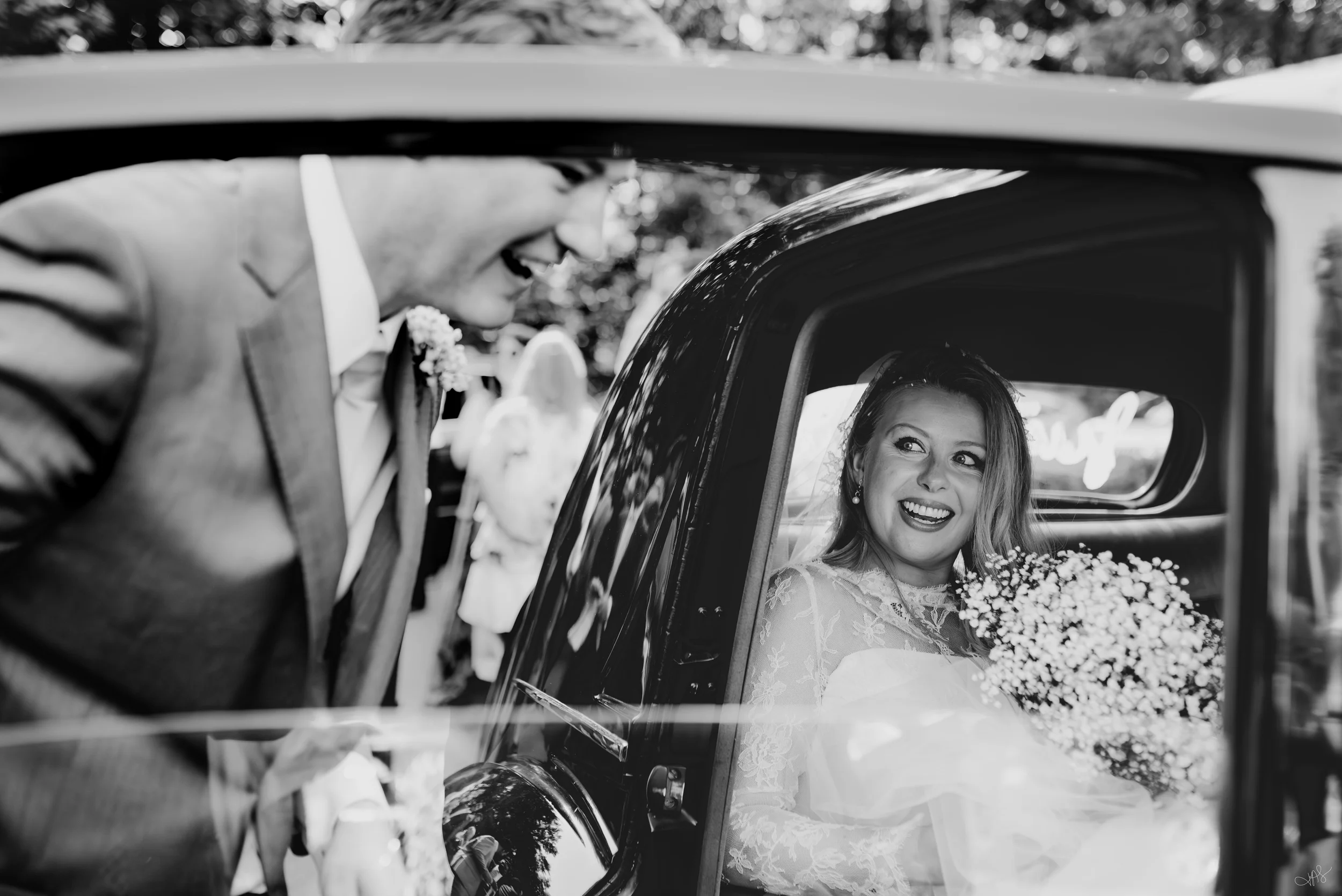 A bride with long hair and earrings sitting inside a car, smiling at the groom. The groom, wearing glasses and a suit, leaning towards her through the car window, smiling back. The photo is in black and white.