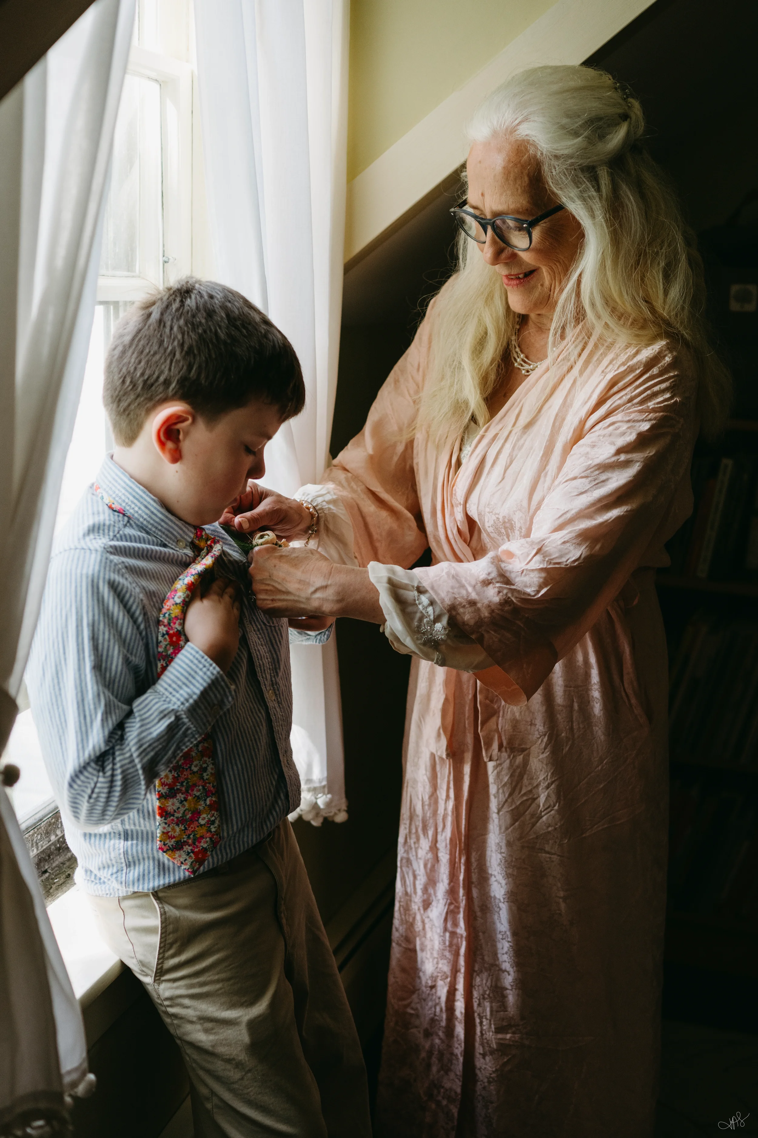 An elderly woman with long, gray hair and glasses is smiling and helping a young boy adjust his clothing or tie near a window with white curtains.