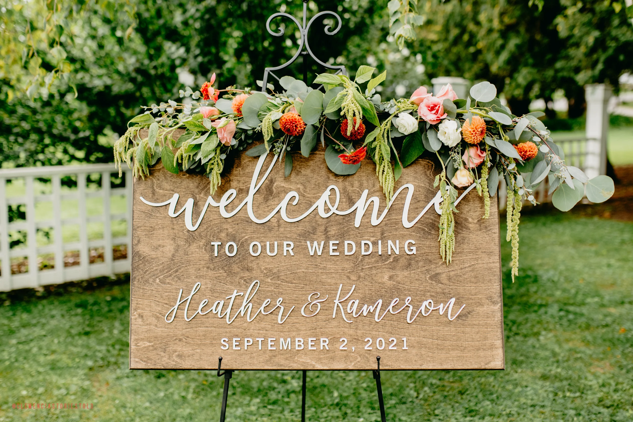 Wooden wedding welcome sign decorated with pink, orange, white flowers, and green foliage outdoors on a grassy area with trees and a white picket fence in the background.