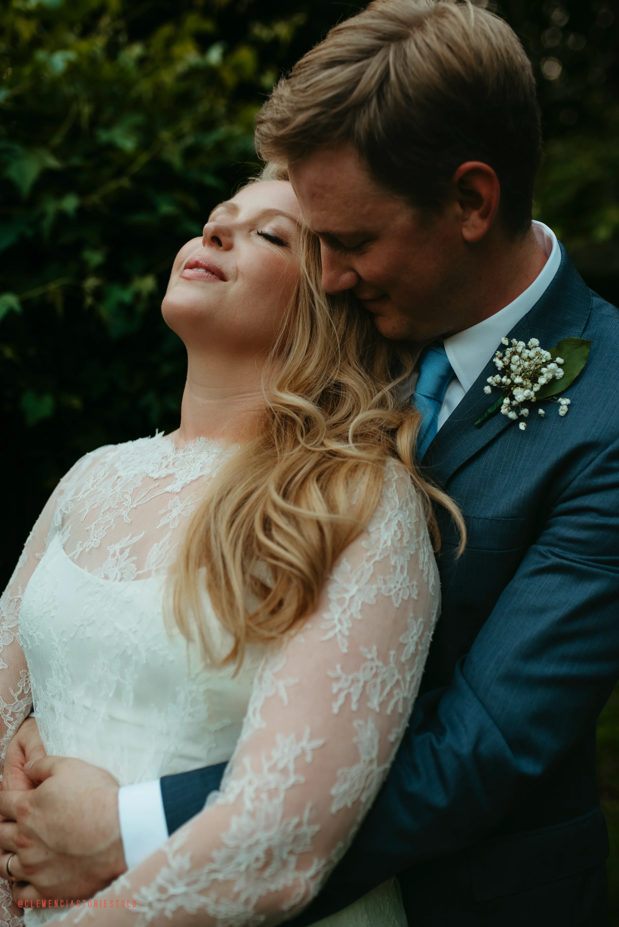 A bride and groom embracing outdoors, with the bride tilting her head back in a joyful, relaxed expression, and the groom leaning into her, dressed in a dark suit with a boutonniere, surrounded by greenery.