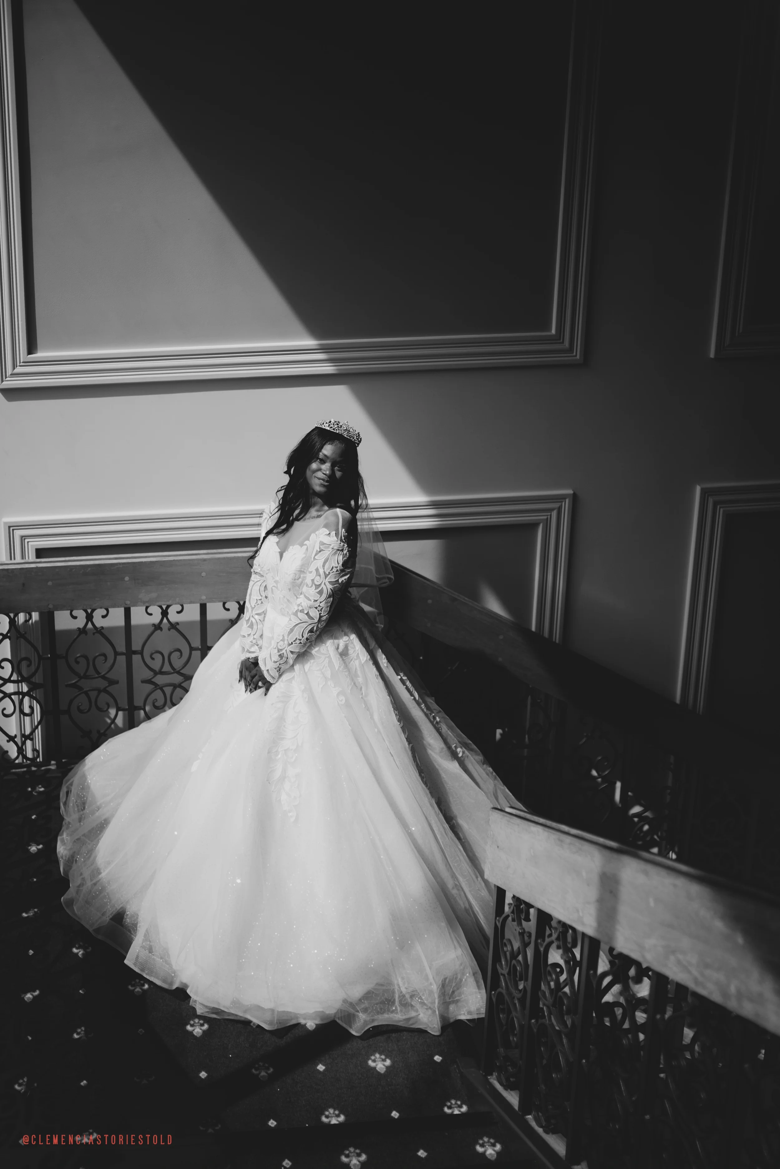 A woman in a wedding dress and tiara standing on a staircase, with a play of light and shadows on the wall behind her.