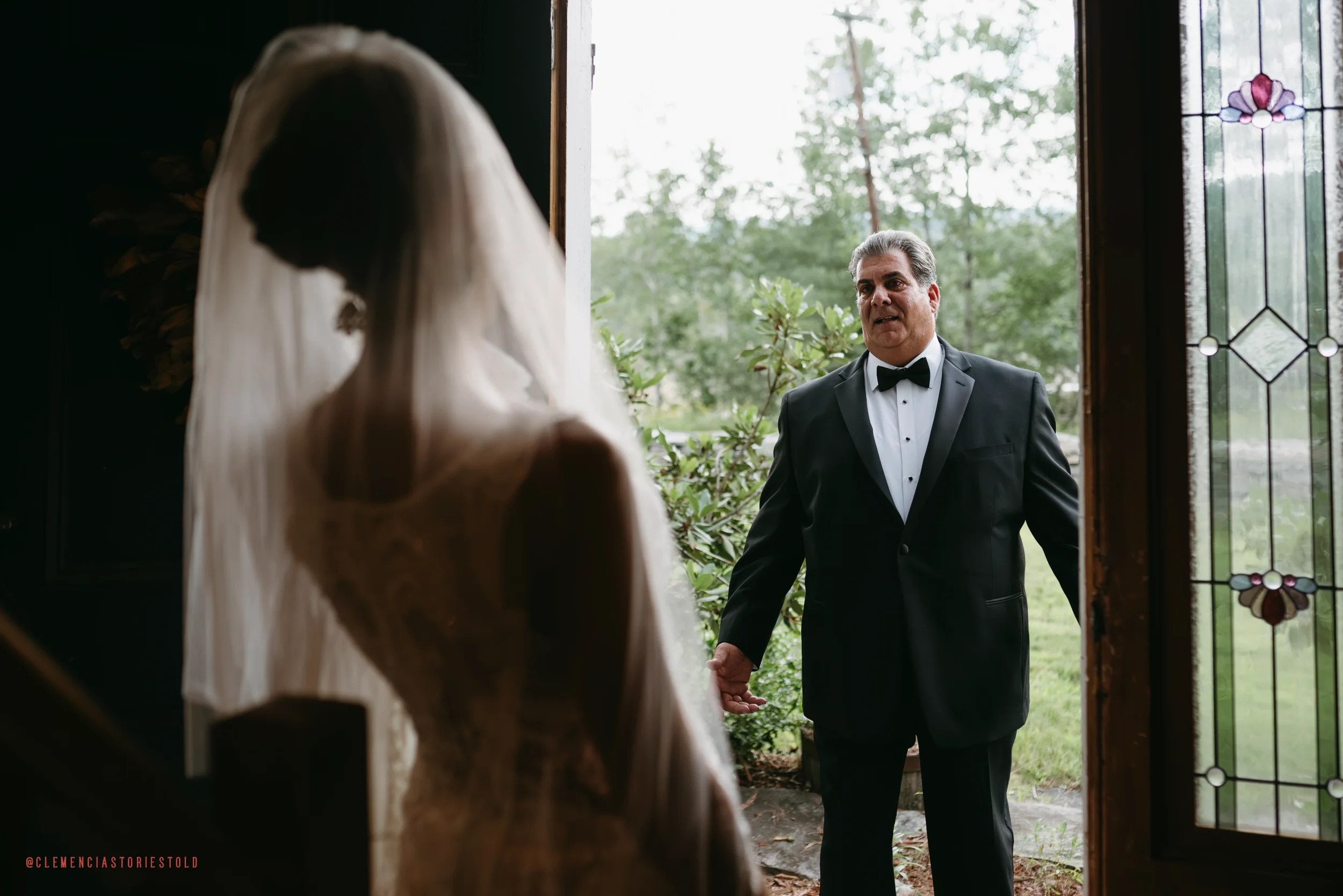 A bride wearing a wedding dress and veil looking at her father, who is standing at the doorway dressed in a tuxedo with a bow tie, during a wedding moment.