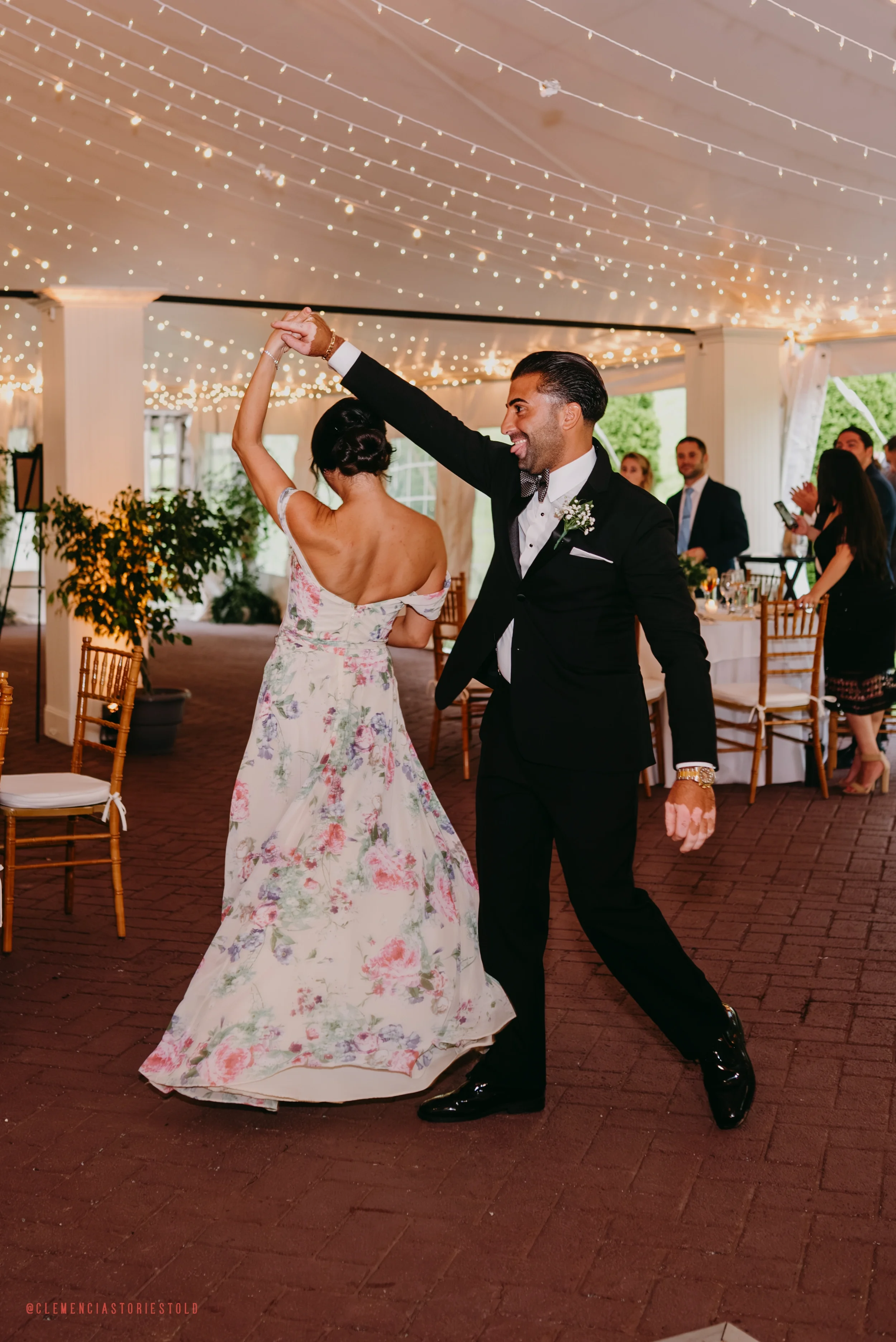A man and woman dancing at a wedding reception under string lights, with seated guests in the background.
