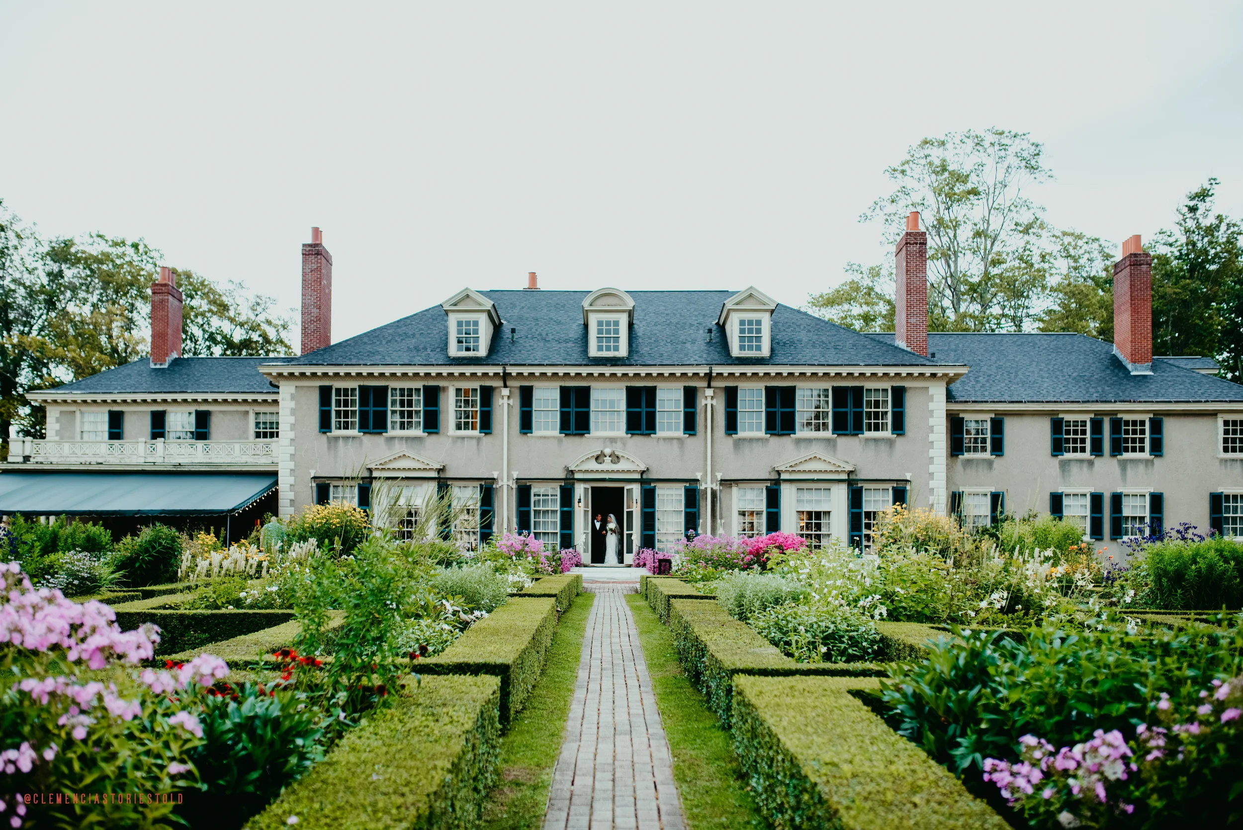 A large, historic white mansion with black shutters and multiple chimneys, surrounded by a manicured garden with colorful flowers and a brick pathway leading to the front door.
