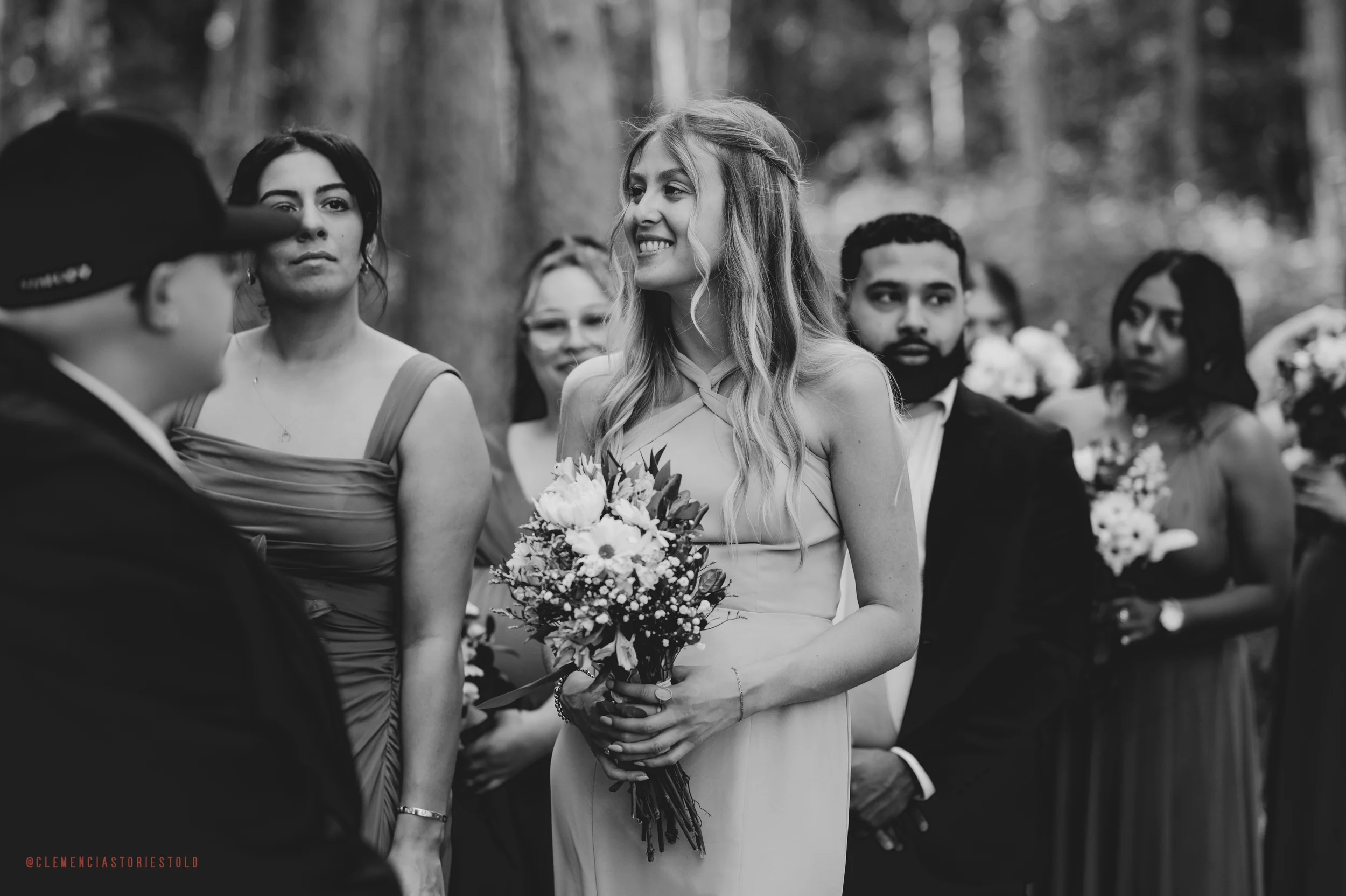 A woman holding a bouquet of flowers smiling during her wedding ceremony outdoors, surrounded by guests in formal attire, in a wooded area.