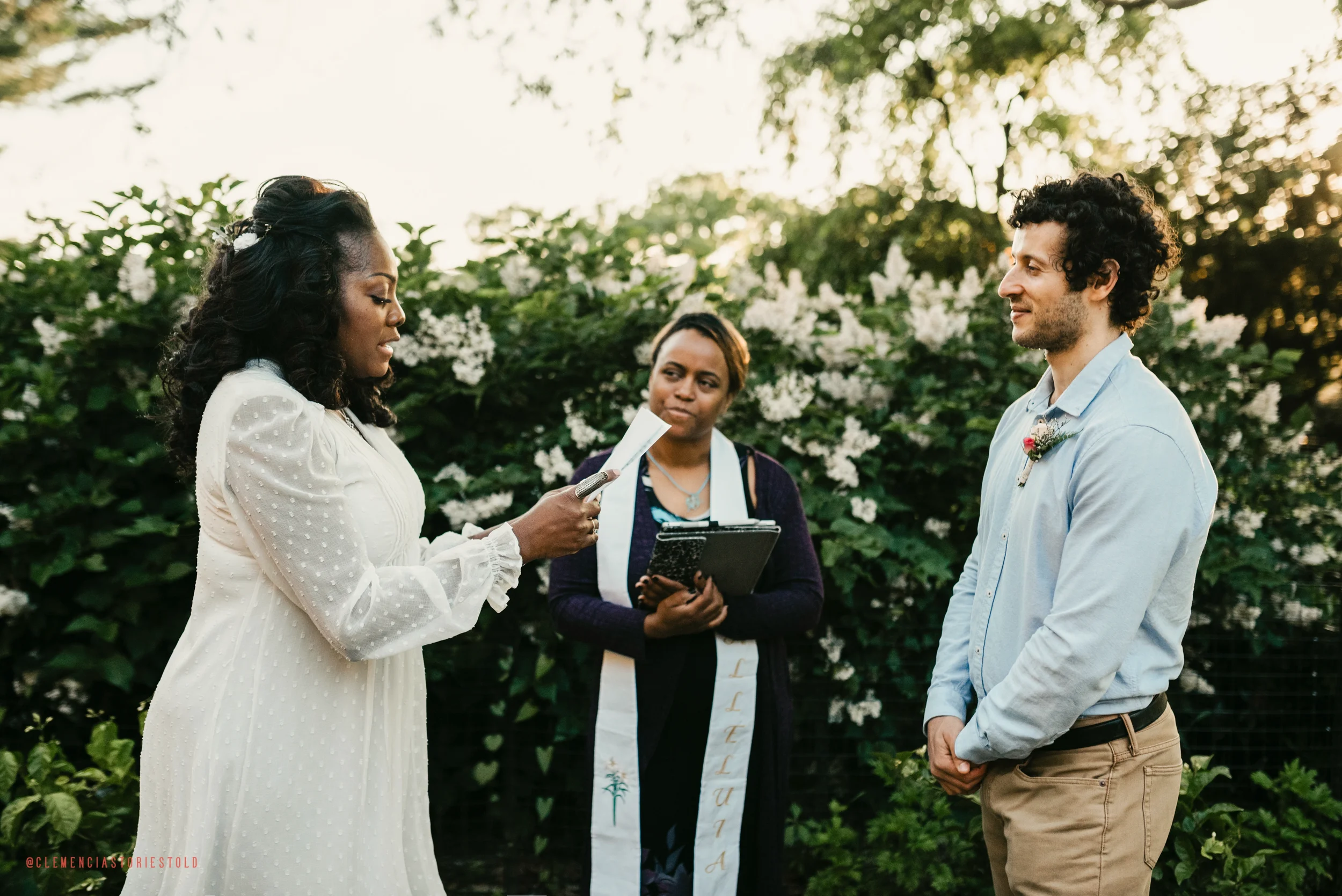 A woman in a white dress officiates a wedding ceremony outdoors, with a man and a woman standing before her. The woman appears to be reading vows or a speech, while the man looks at her with a smile. The ceremony is set against lush greenery and whit