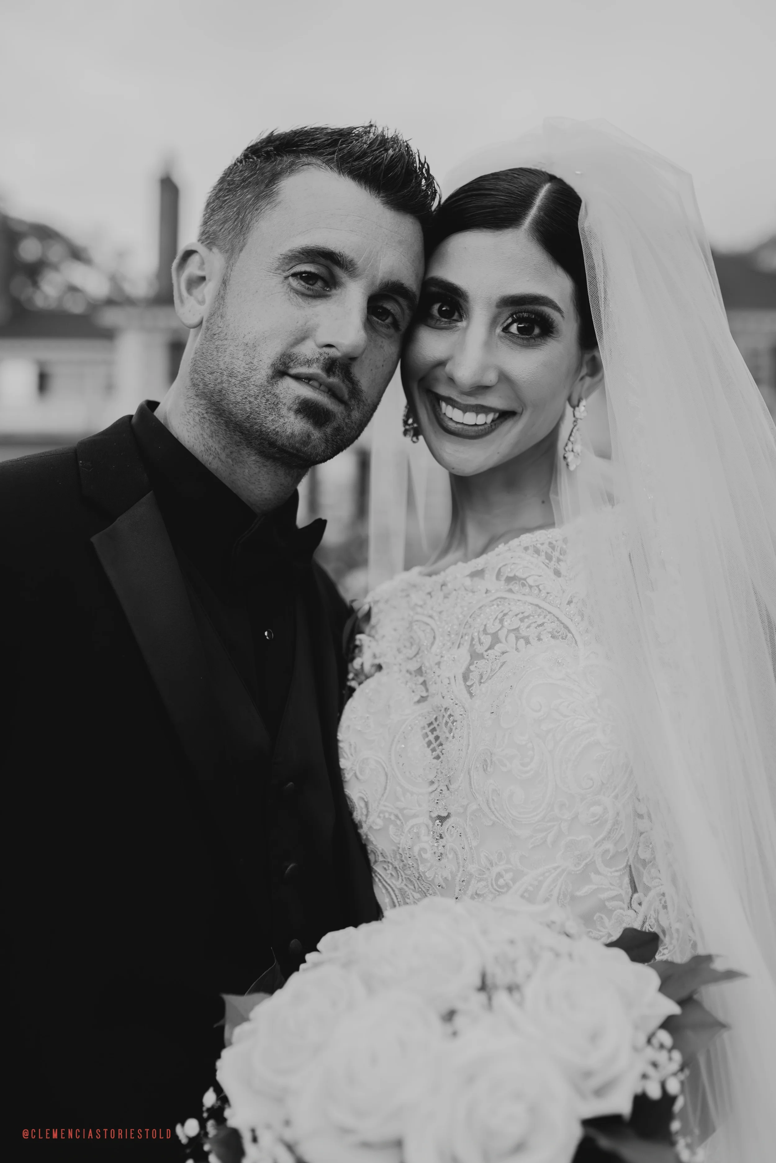 Black-and-white photo of a smiling bride and groom close together, the bride wearing a lace wedding dress and veil, holding a bouquet of roses, with buildings in the background.
