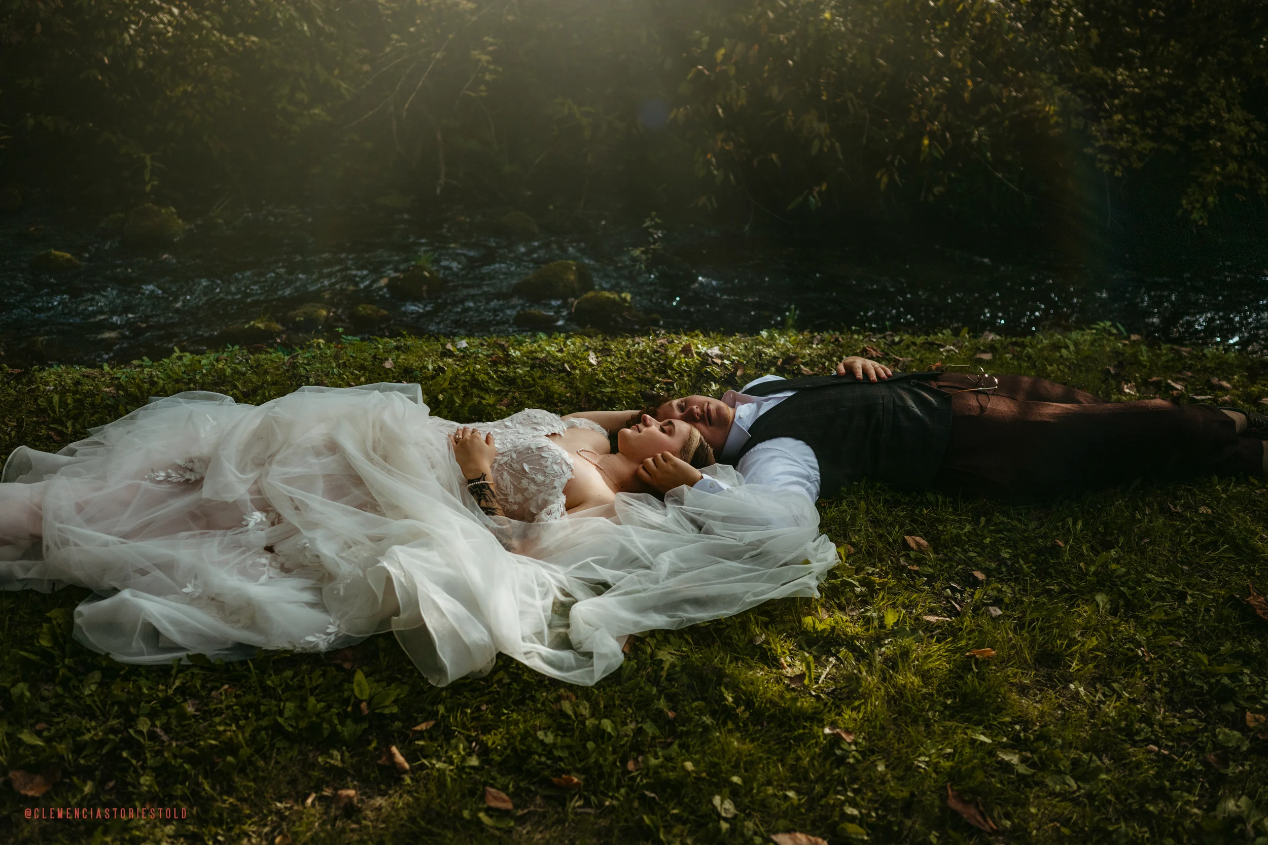 A bride and groom lying on grass near a river, holding hands and resting with eyes closed, during a wedding photoshoot in a natural setting.