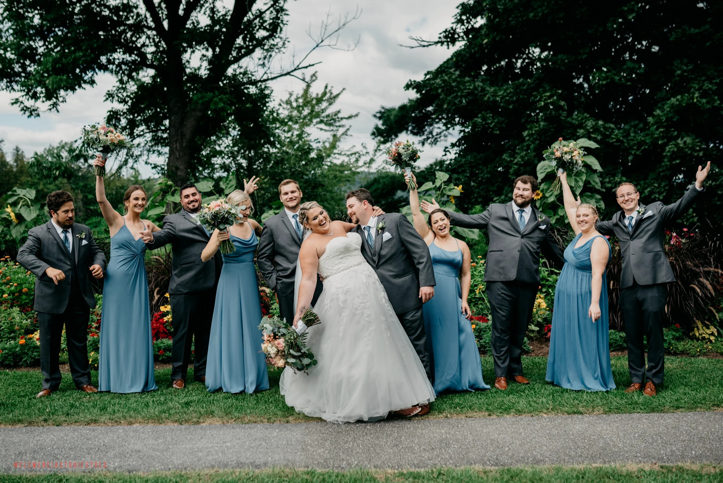 A group of people dressed in wedding attire posing outdoors on a cloudy day, with some holding bouquets and others celebrating with raised hands.