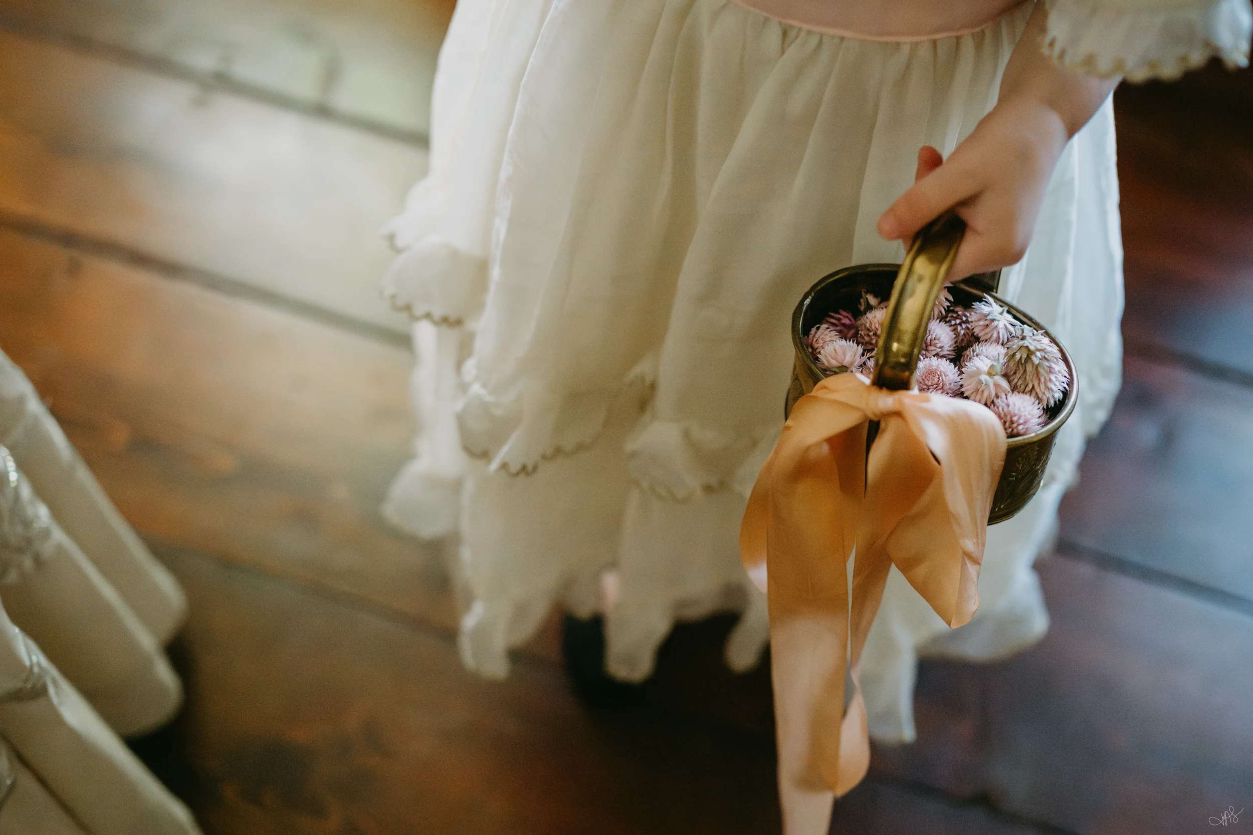 A person in a vintage cream-colored dress is holding a small bouquet of pink flowers inside a ceramic or metal container with a gold handle.