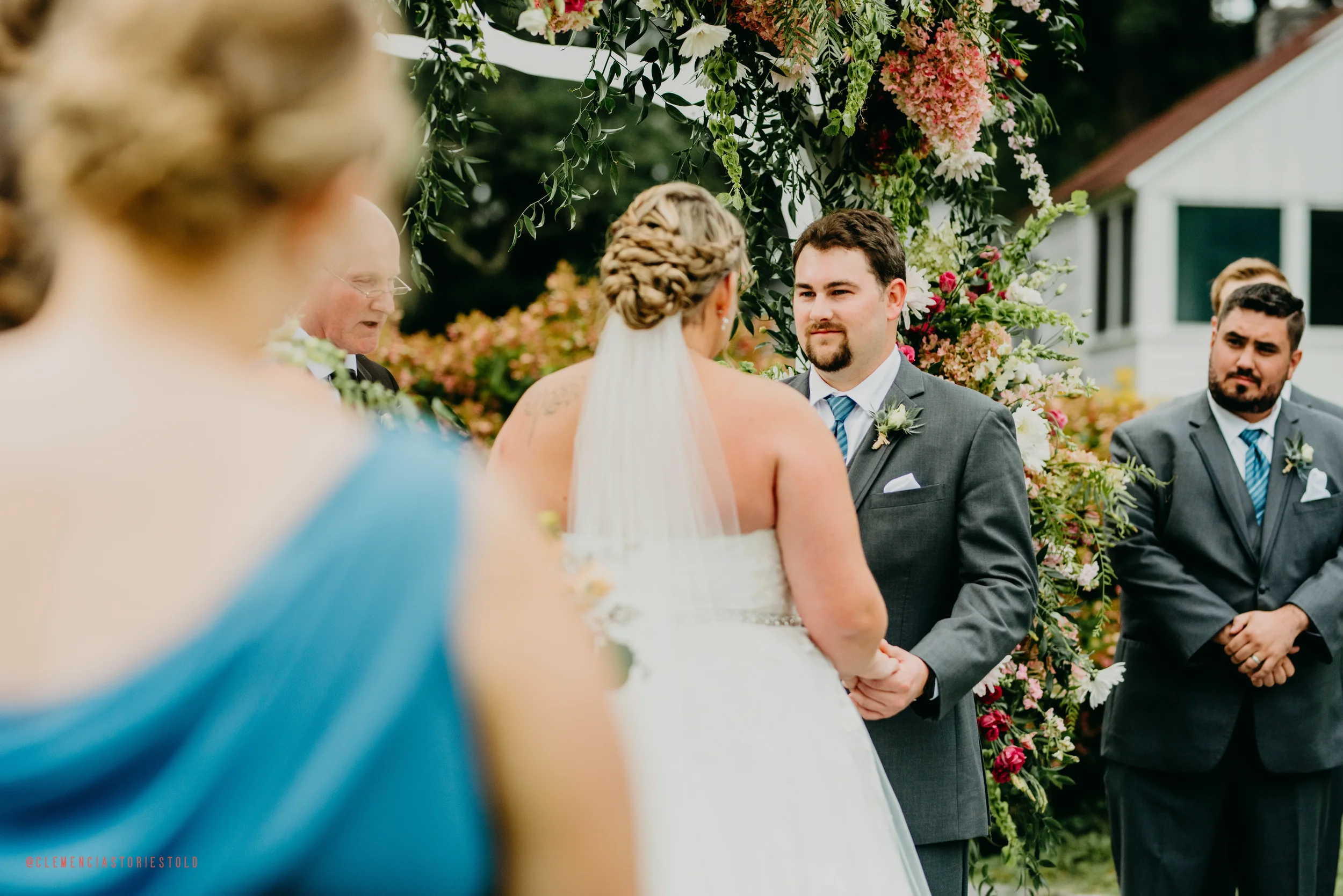 A wedding ceremony outdoors with the bride and groom holding hands and facing each other, surrounded by floral decorations, with an officiant and groomsman visible in the background.