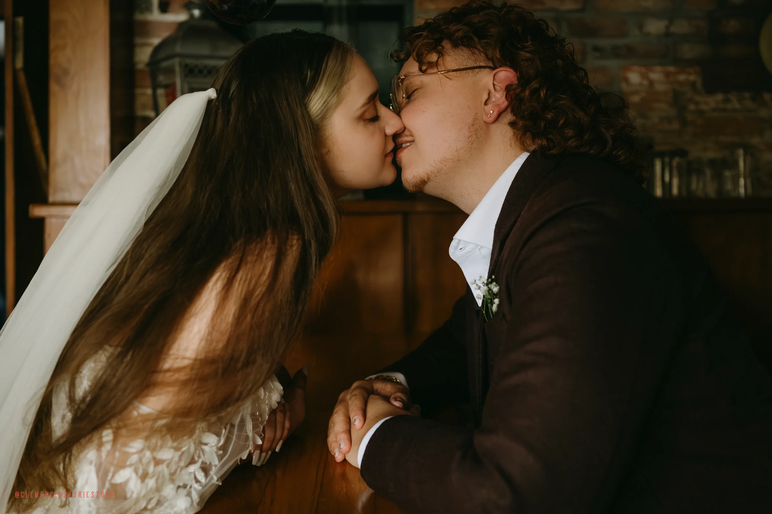 A couple sharing a kiss at their wedding, with the bride wearing a white veil and dress, and the groom wearing glasses and a black tuxedo, sitting at a wooden table in a cozy, rustic setting.