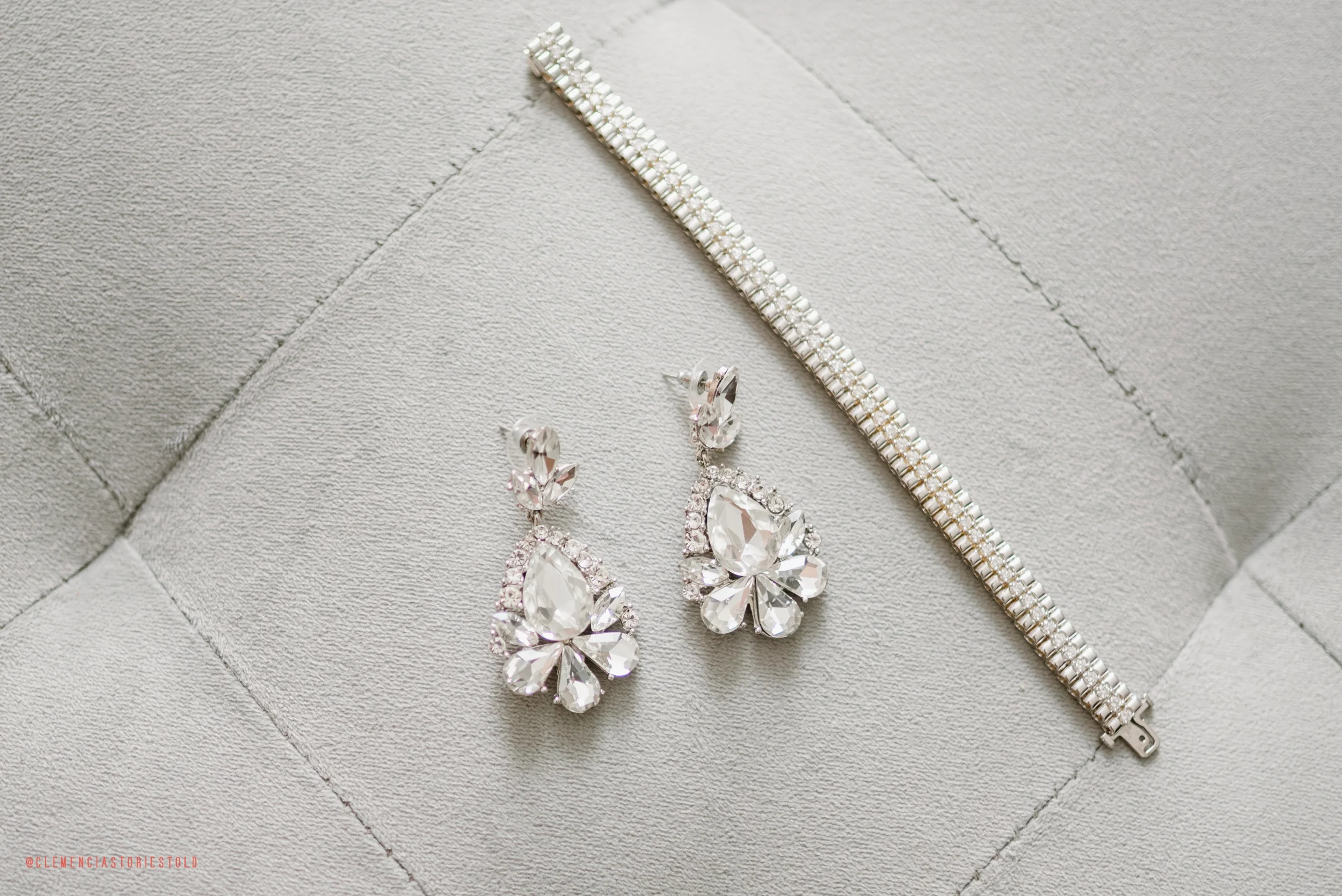 Pair of silver earrings with large teardrop-shaped crystals and smaller surrounding crystals, and a silver bracelet with rectangular links and embedded smaller crystals on a light gray textured surface.