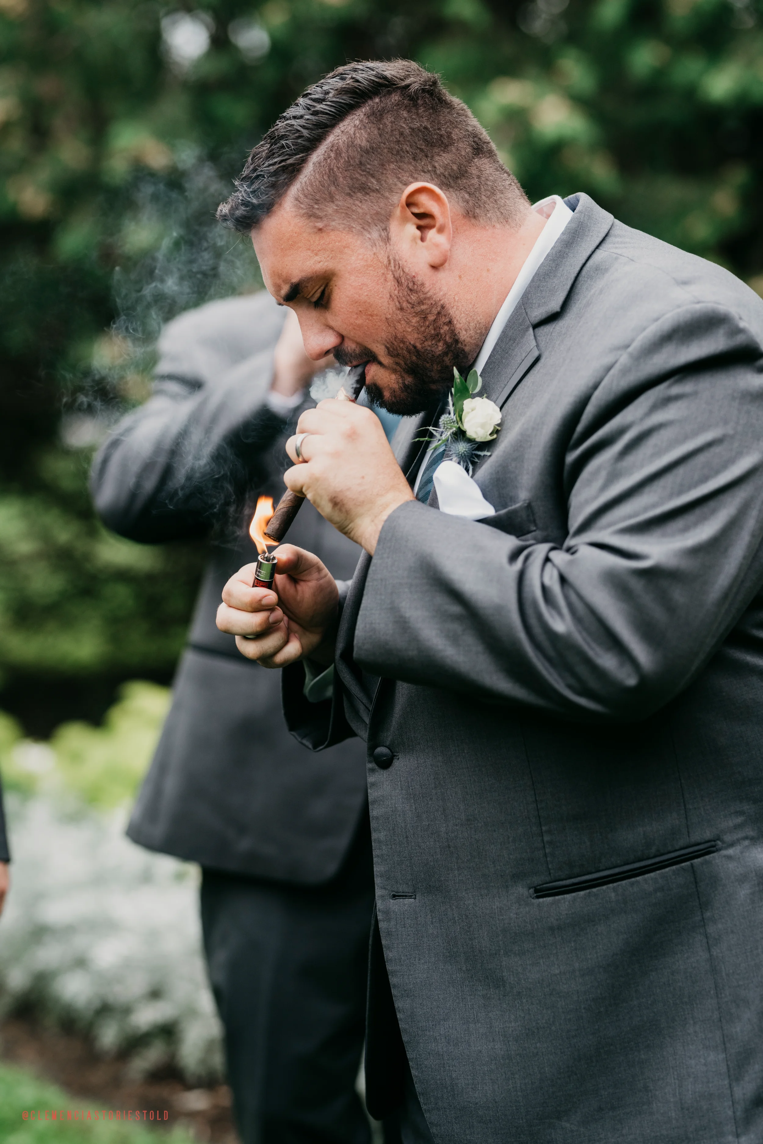A man in a gray suit lighting a cigar with a lighter at an outdoor event, with blurred greenery in the background.