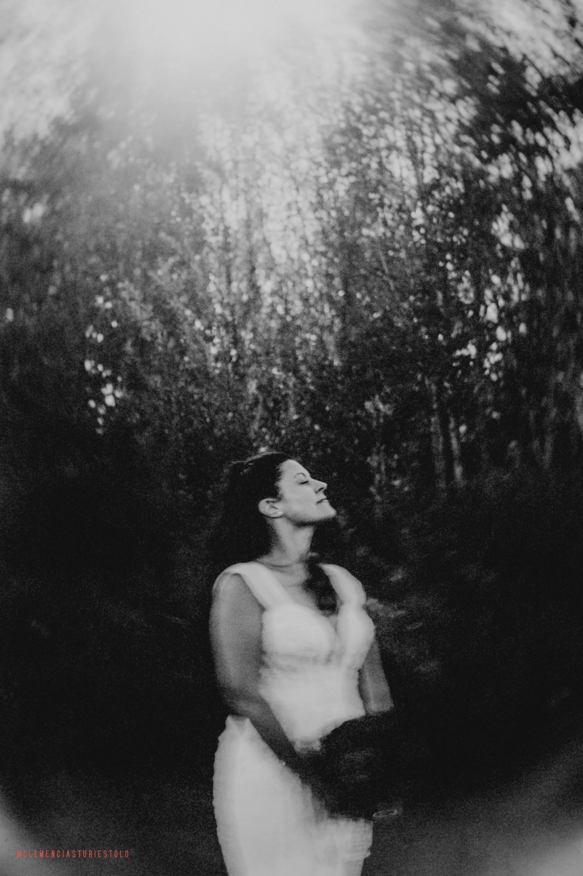 A black and white photo of a woman with long dark hair in a dress standing in a body of water with trees in the background. She has her eyes closed and her face turned slightly upward, with sunlight filtering through the trees.
