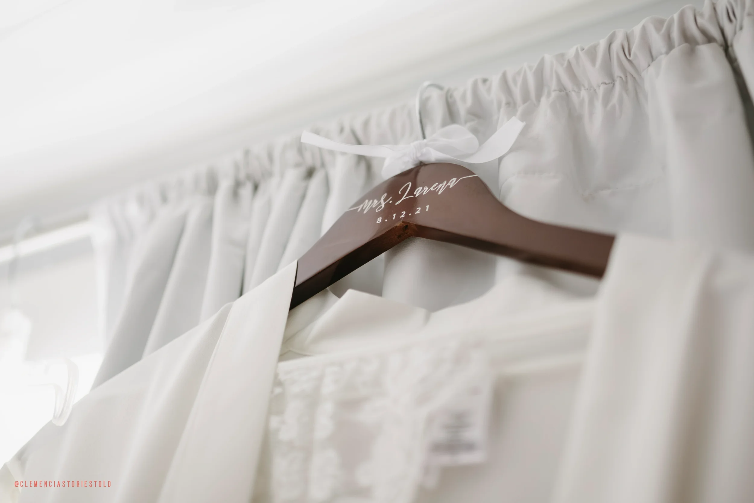 A wooden hanger with embroidered text reading 'Mrs. Lareva 8.12.21' hanging on a white curtain rod.
