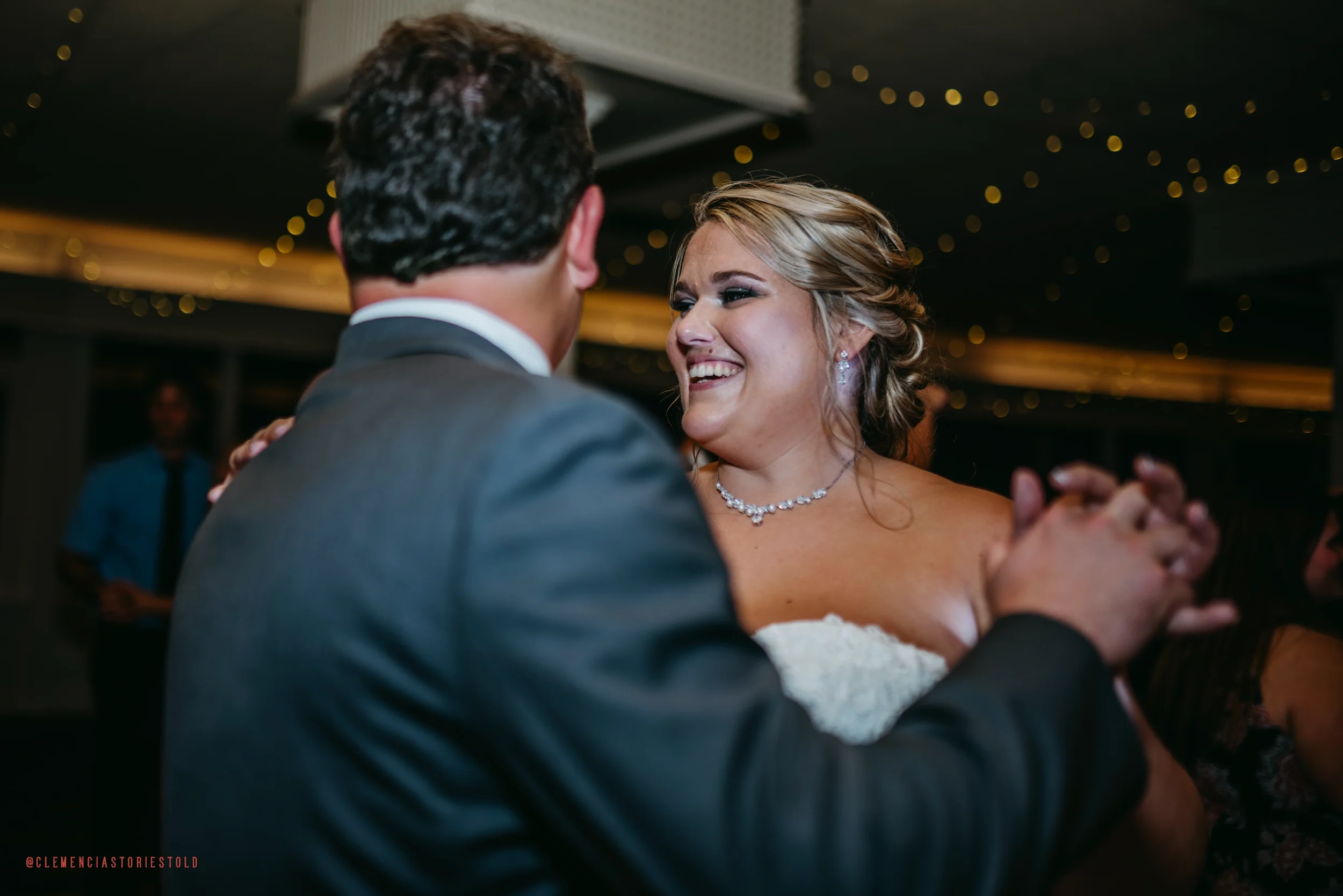 A bride and groom dancing at their wedding reception, smiling and looking at each other, with warm celebratory lighting in the background.