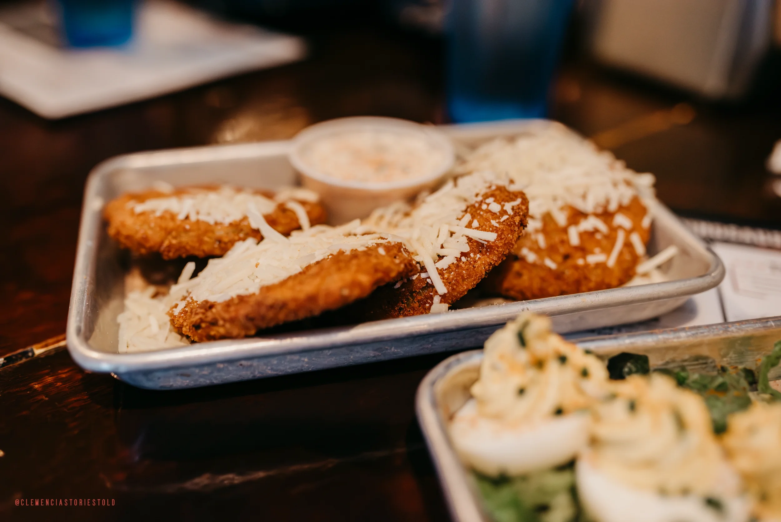 Fried chicken topped with shredded cheese served with a side of dipping sauce on a metal tray.