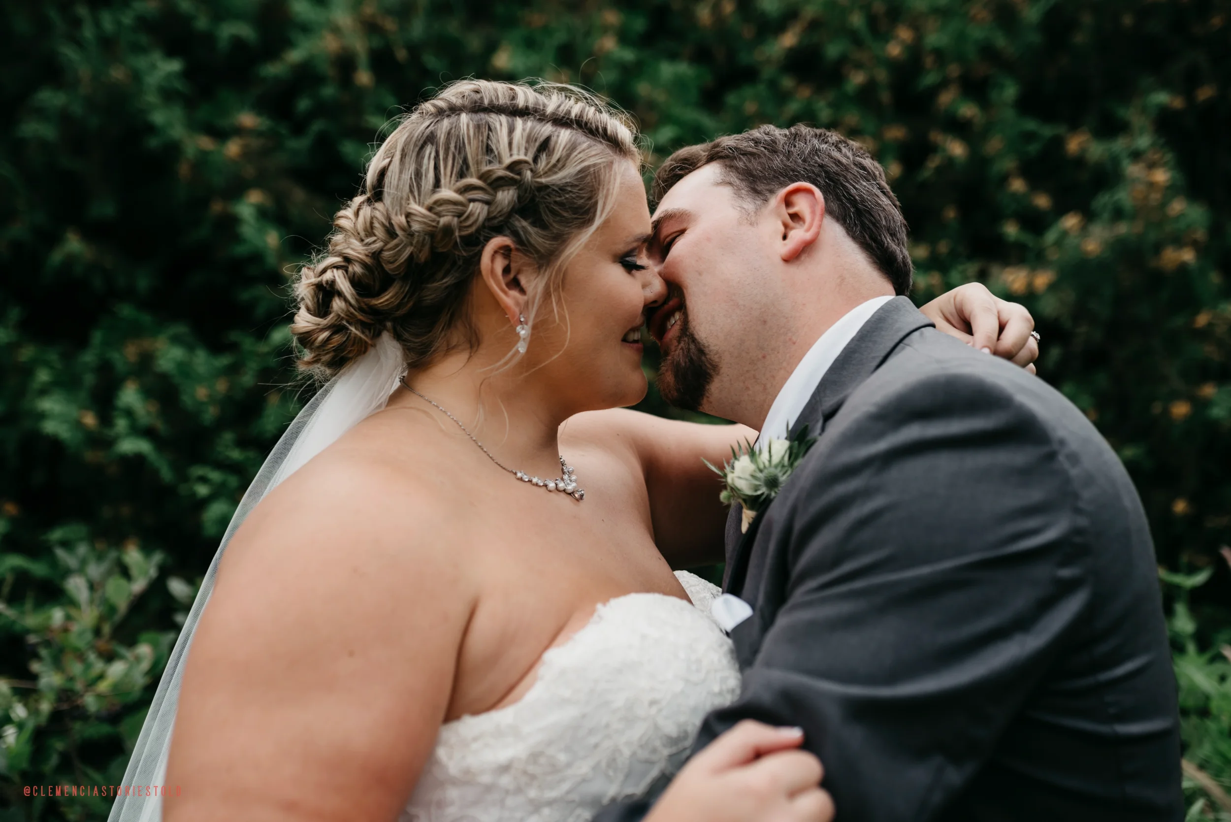A bride and groom are close, touching foreheads and smiling outdoors with green foliage in the background.